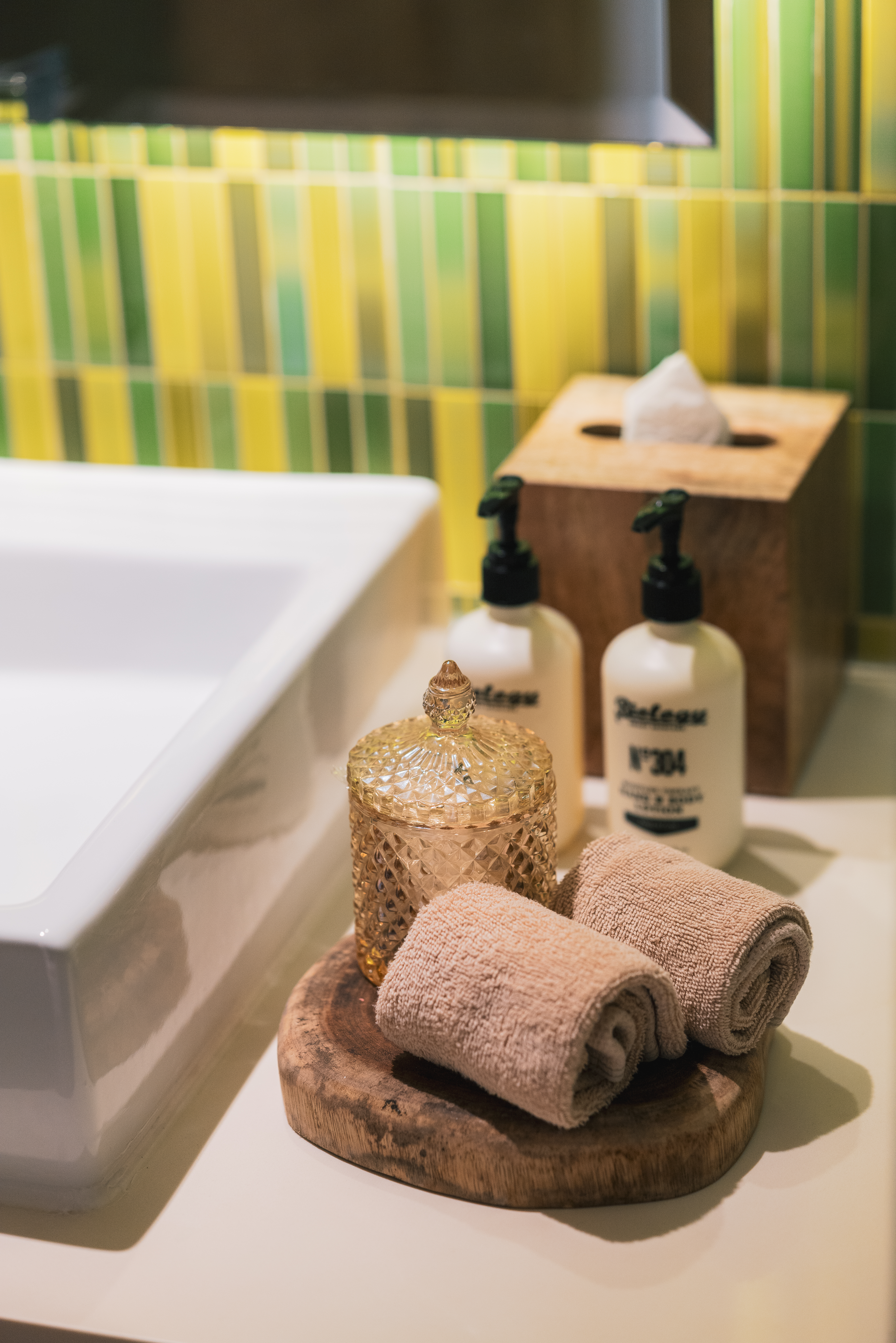 Bathroom counter with white sink, soap, rolled towels, and yellow-green tiles.