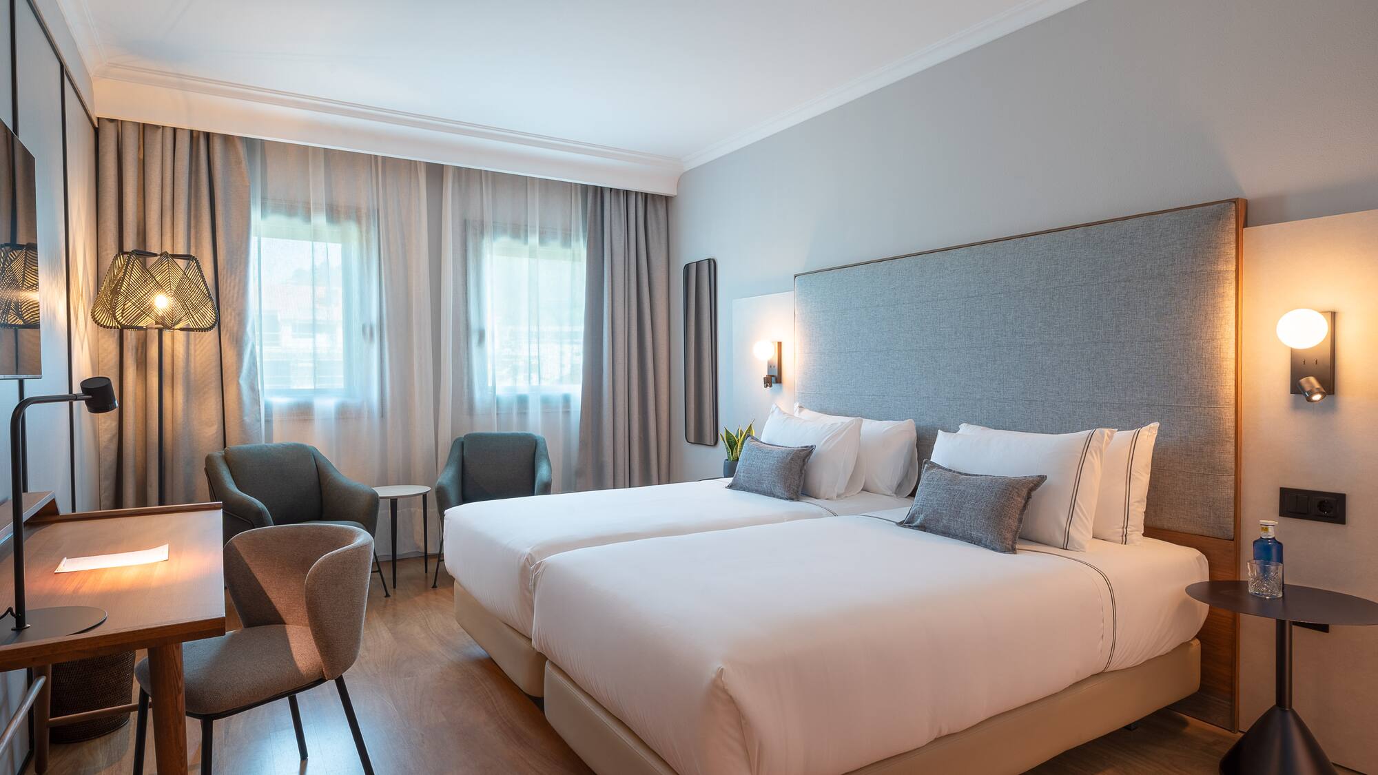 Modern hotel room with twin beds, desk, armchairs, and wood floor.