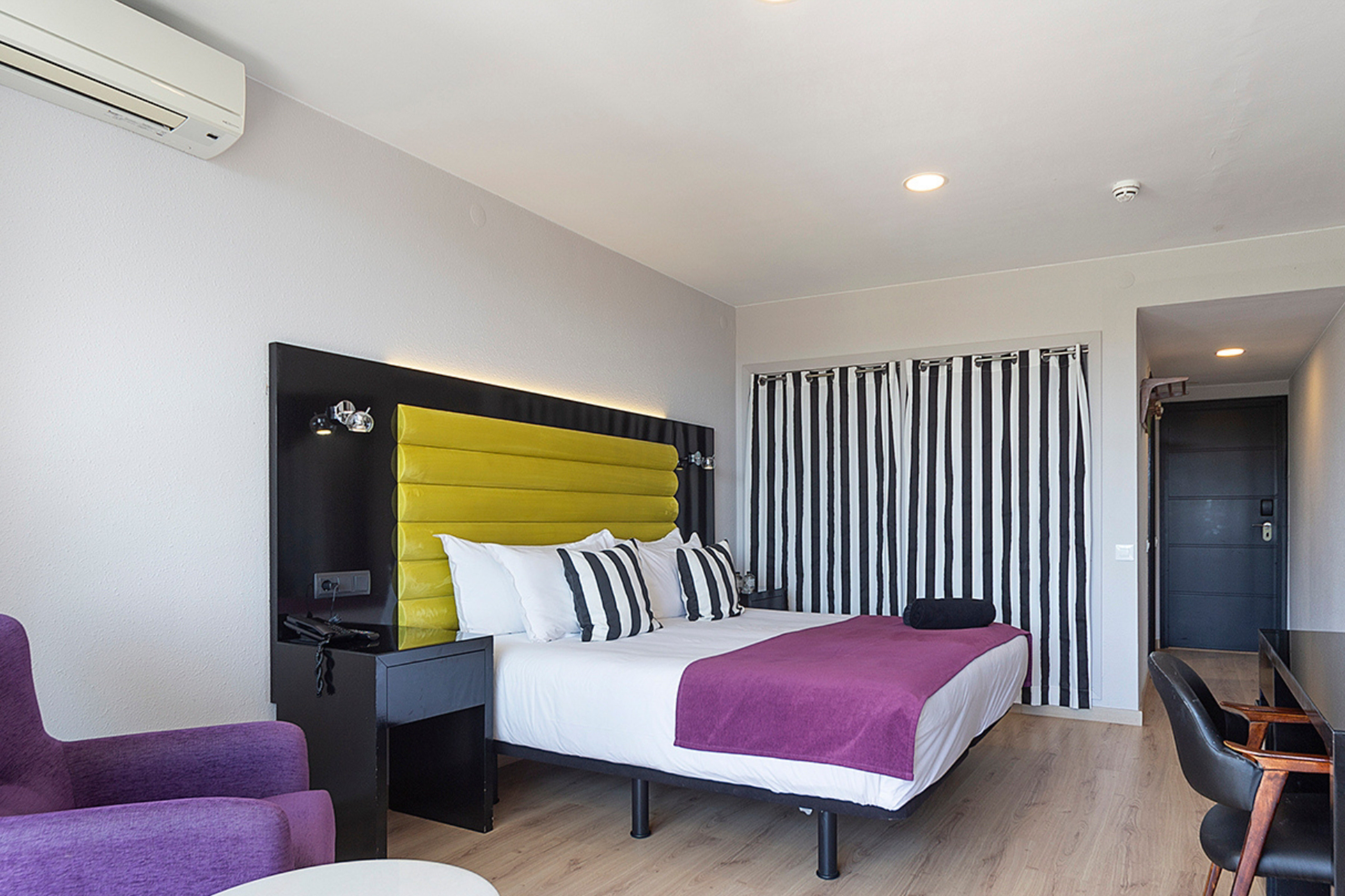 Modern hotel bedroom with yellow headboard, purple bed, and black striped curtains.