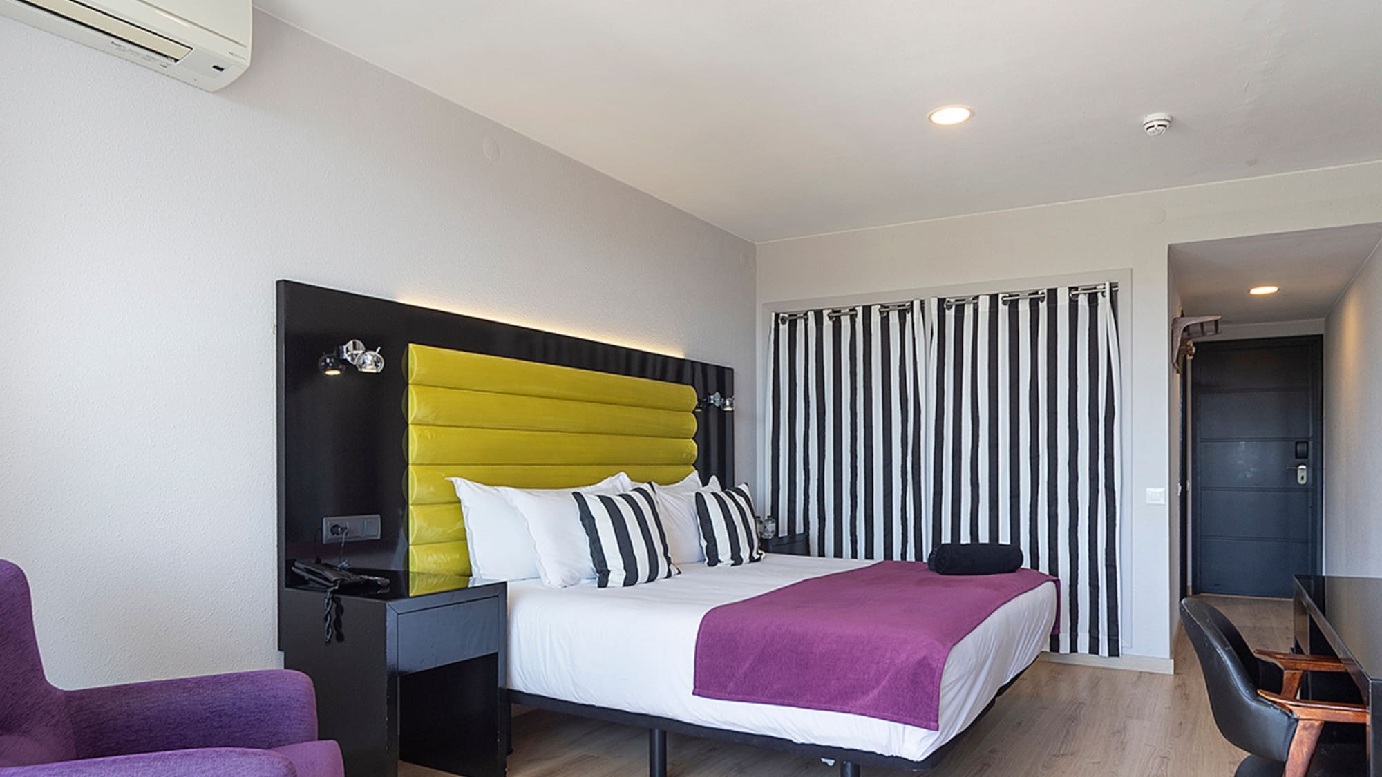Modern hotel bedroom with yellow headboard, purple bed, and black striped curtains.