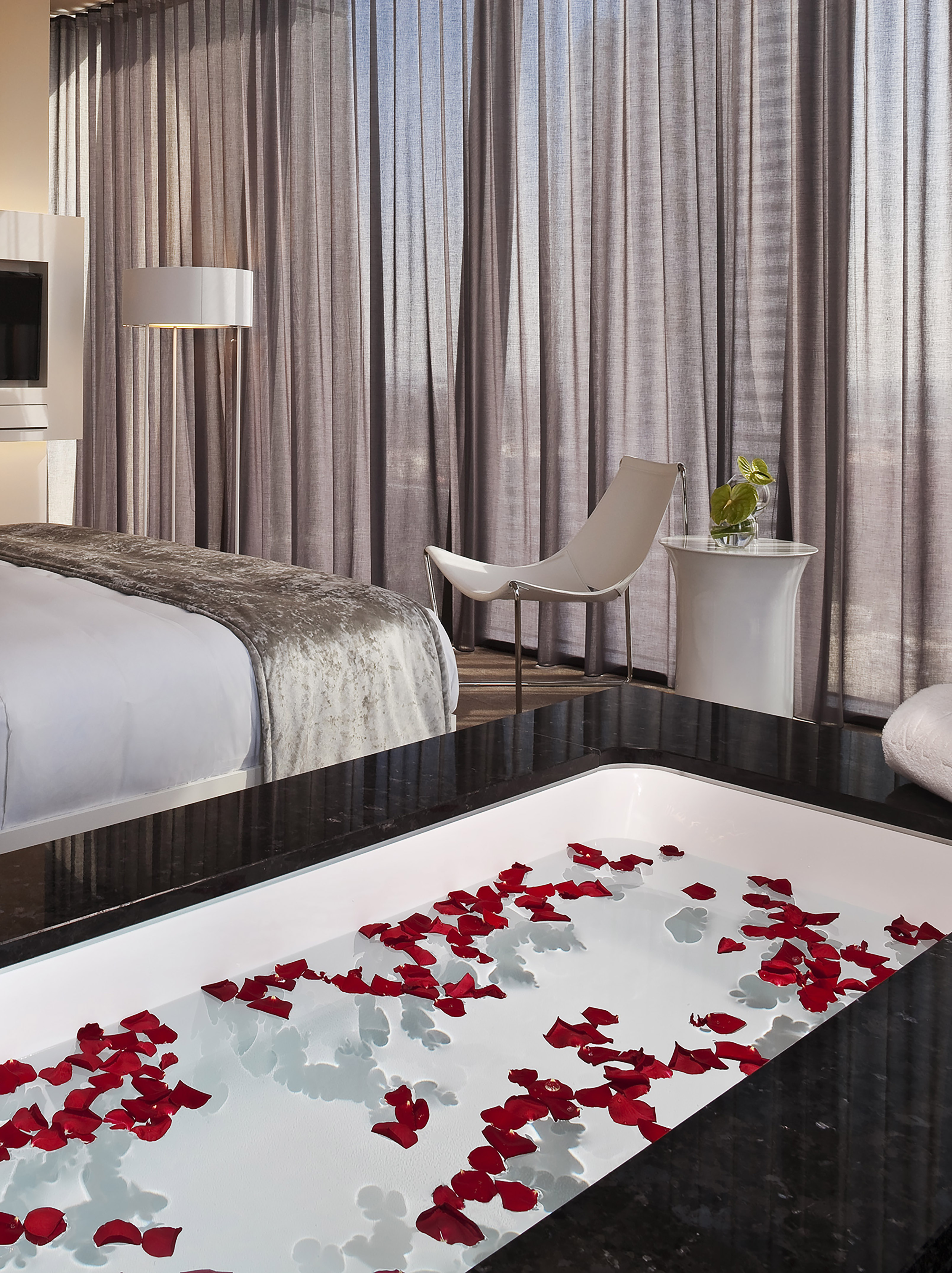 Luxury hotel suite with rose petal bath, modern bed, and sheer curtains.