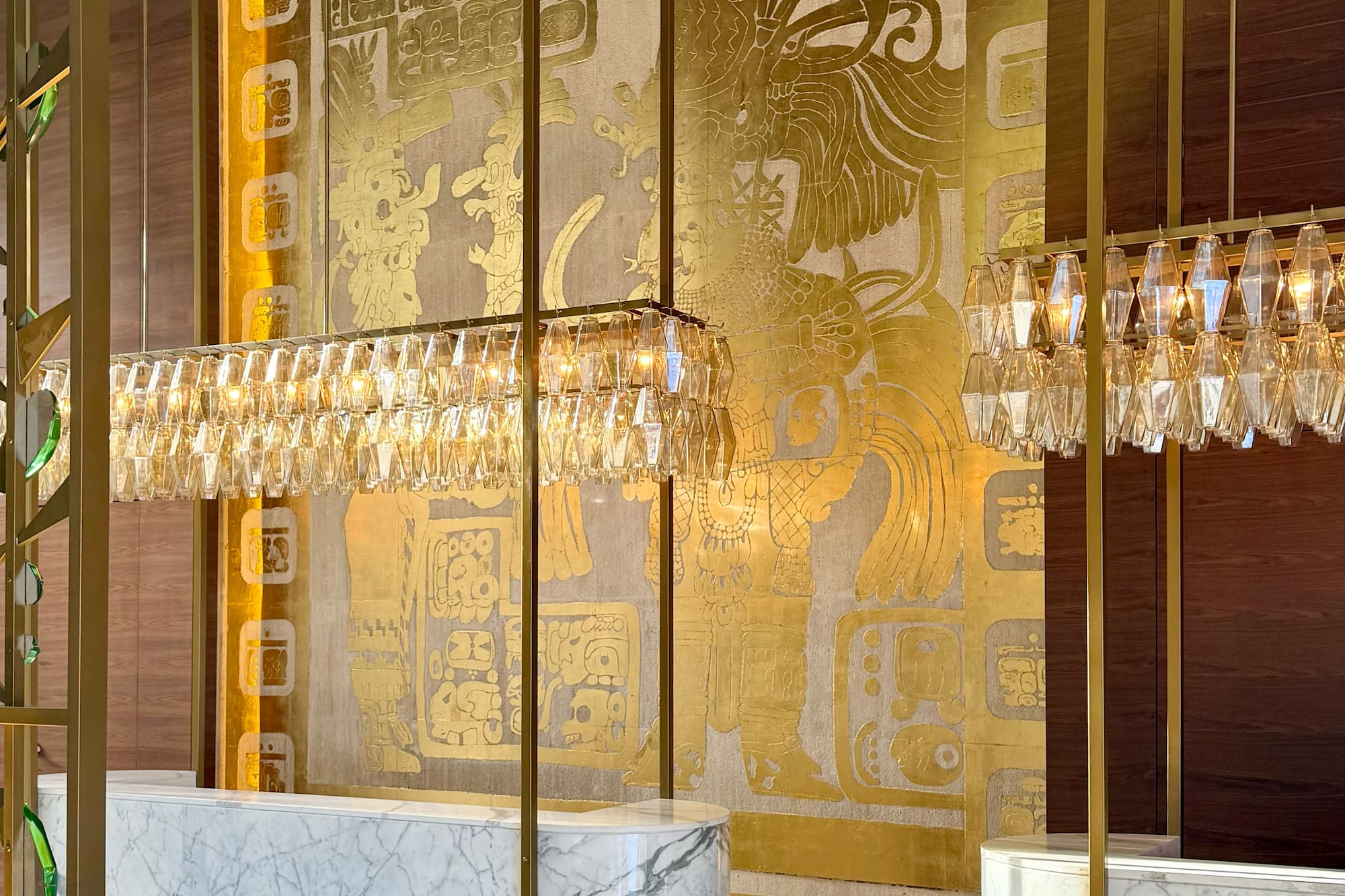 Luxury hotel lobby: marble reception, crystal chandeliers, gold Mayan mural.