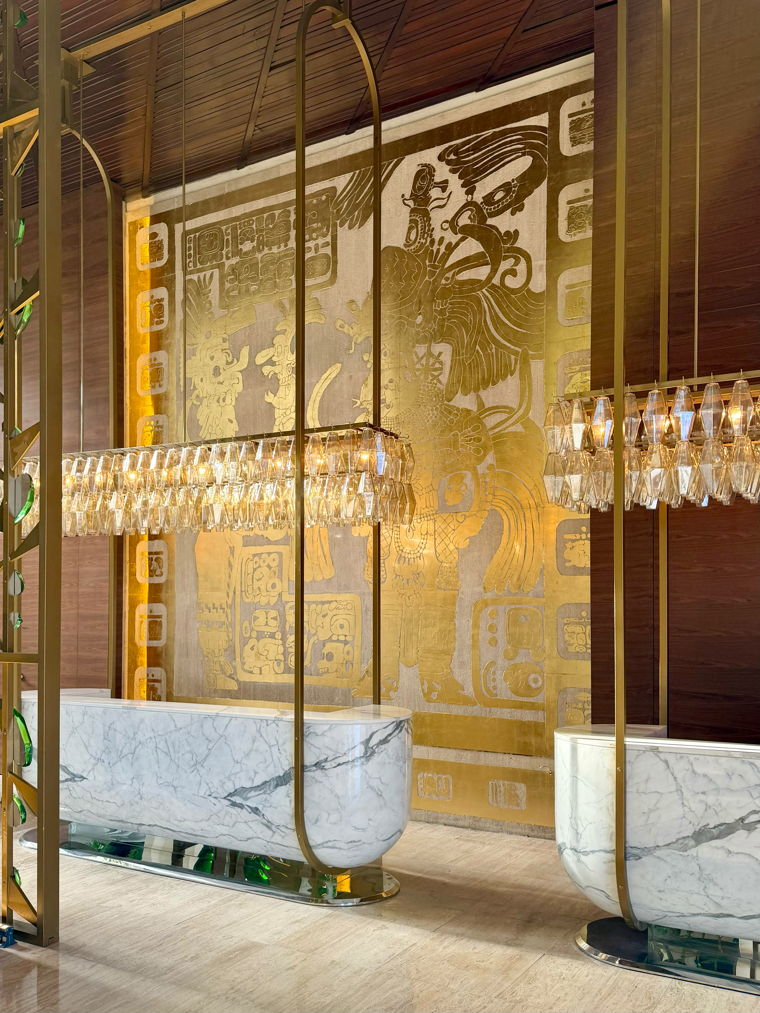 Luxury hotel lobby: marble reception, crystal chandeliers, gold Mayan mural.