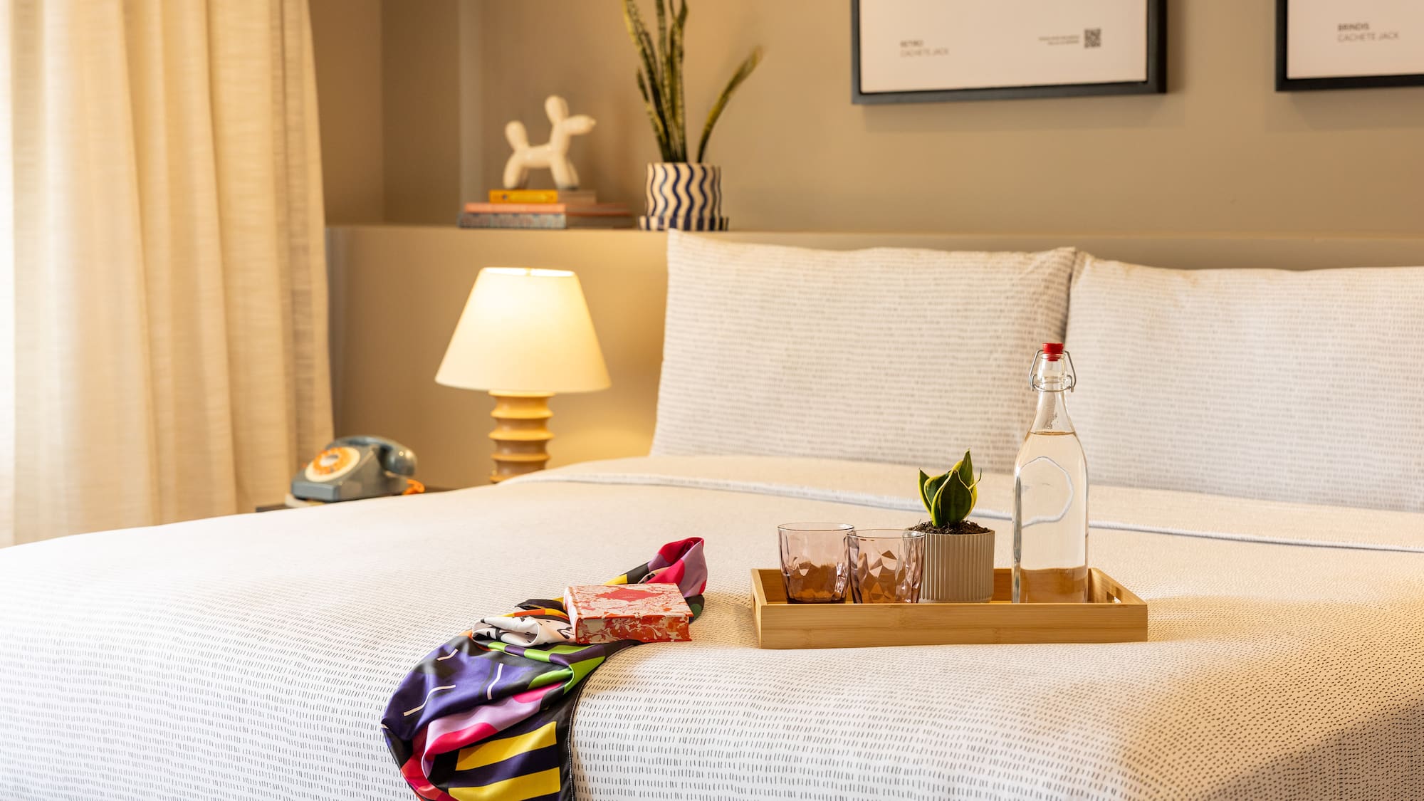 Cama de hotel con bandeja de madera, botella de agua, vasos, plantas y arte mural.