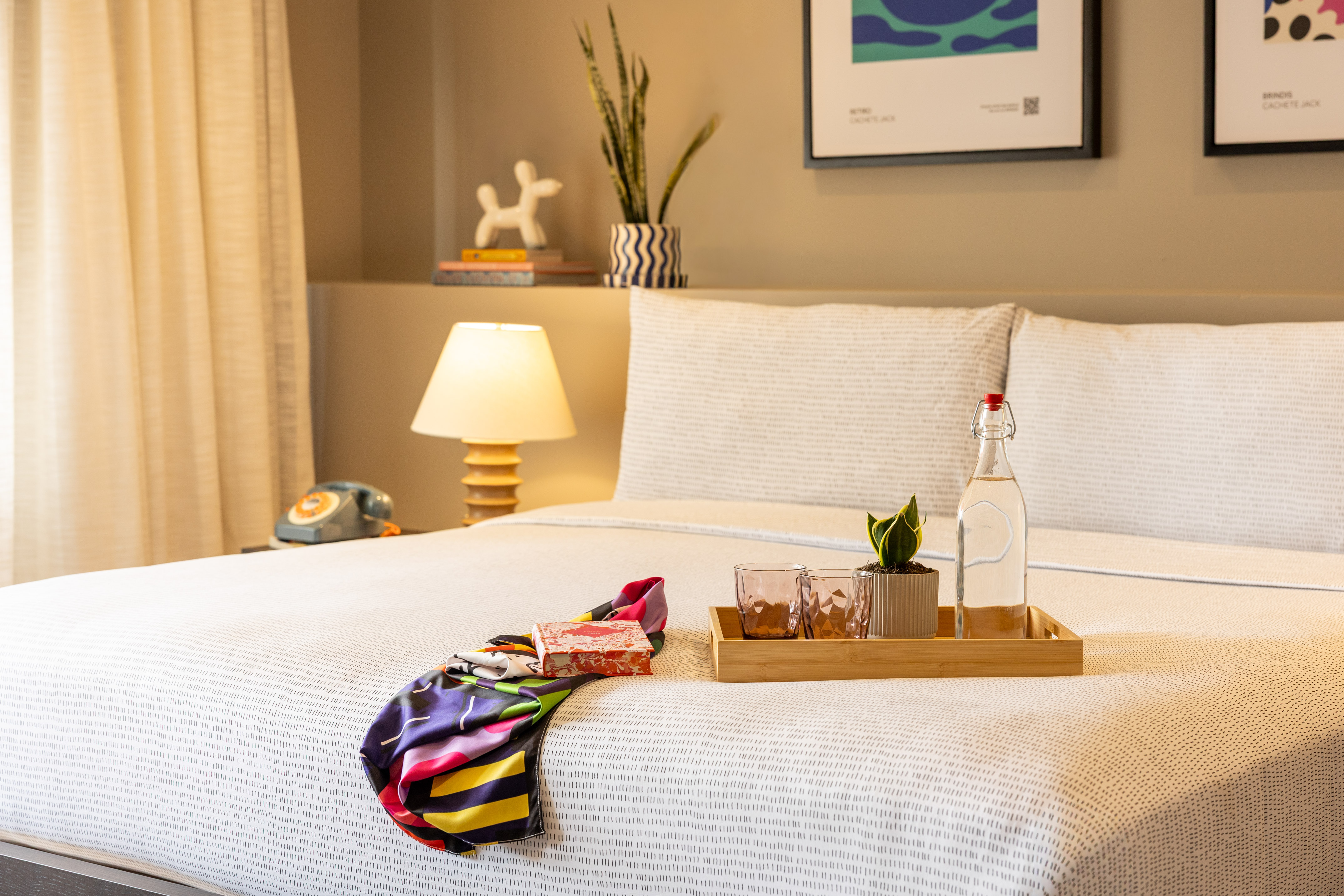 Hotel bed with patterned duvet, colorful scarf, water bottle, and bedside decor.