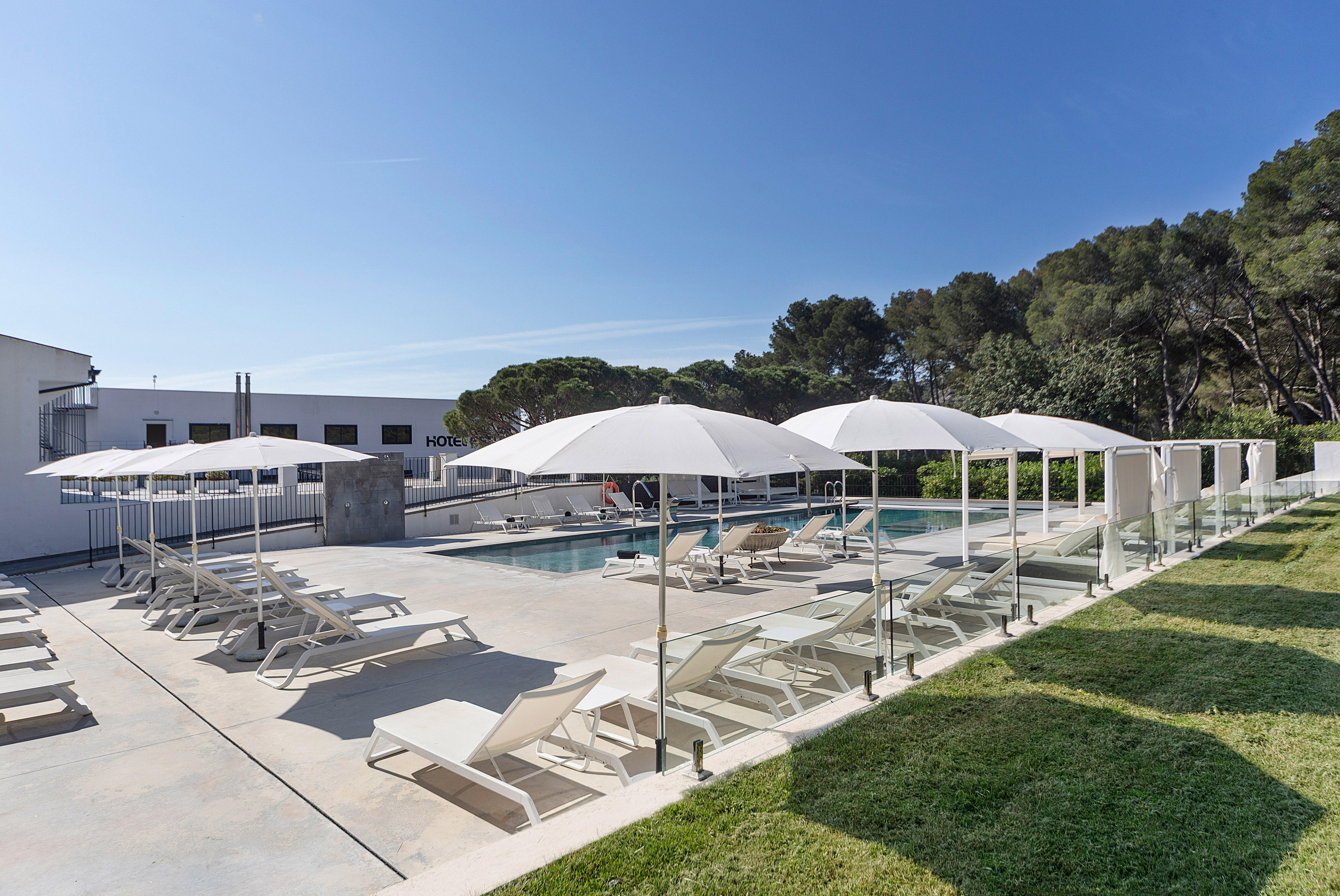 Sunny hotel pool area with white sunbeds, umbrellas, and green grass.