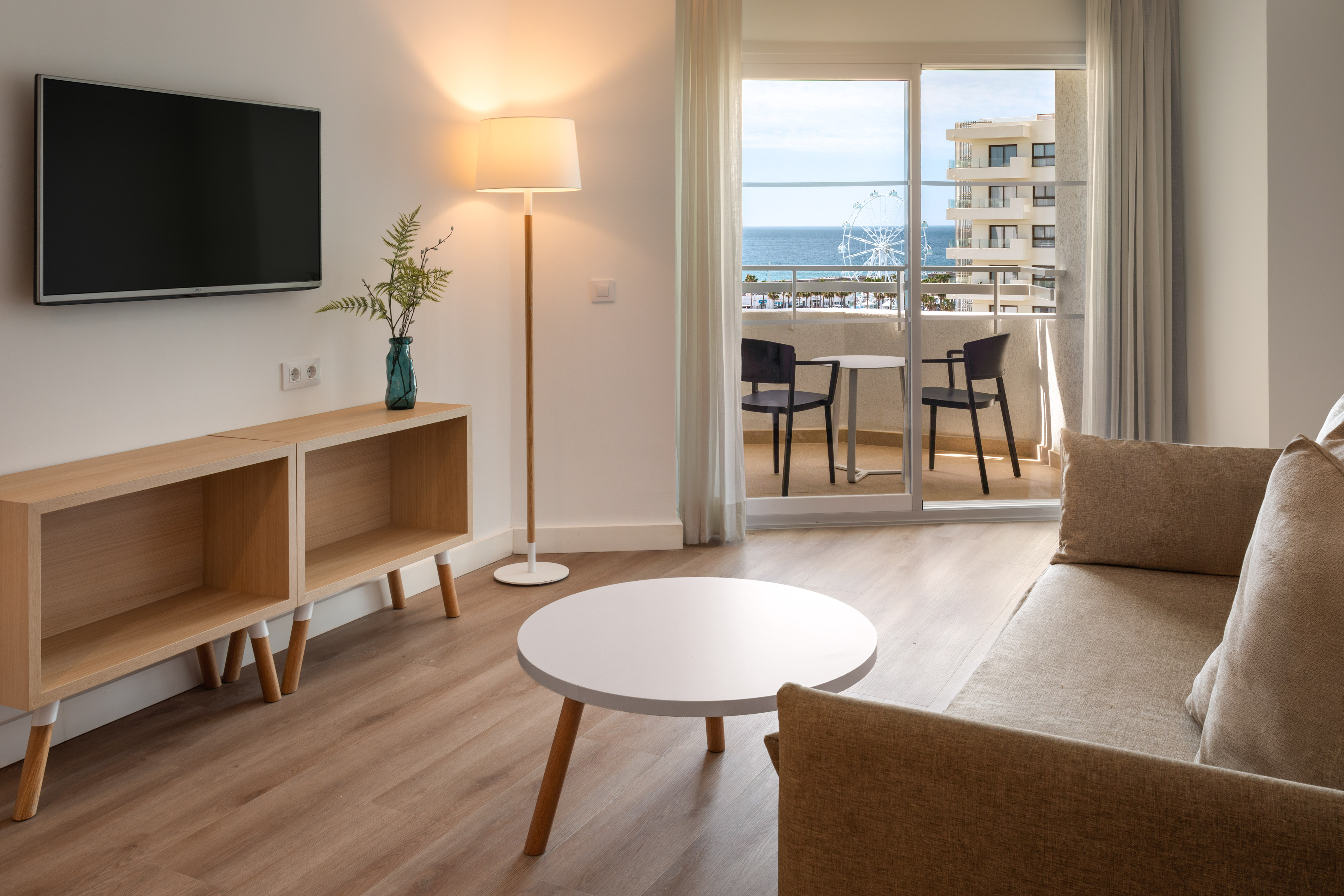 Hotel suite living room with ocean view balcony, TV, sofa, and Ferris wheel.