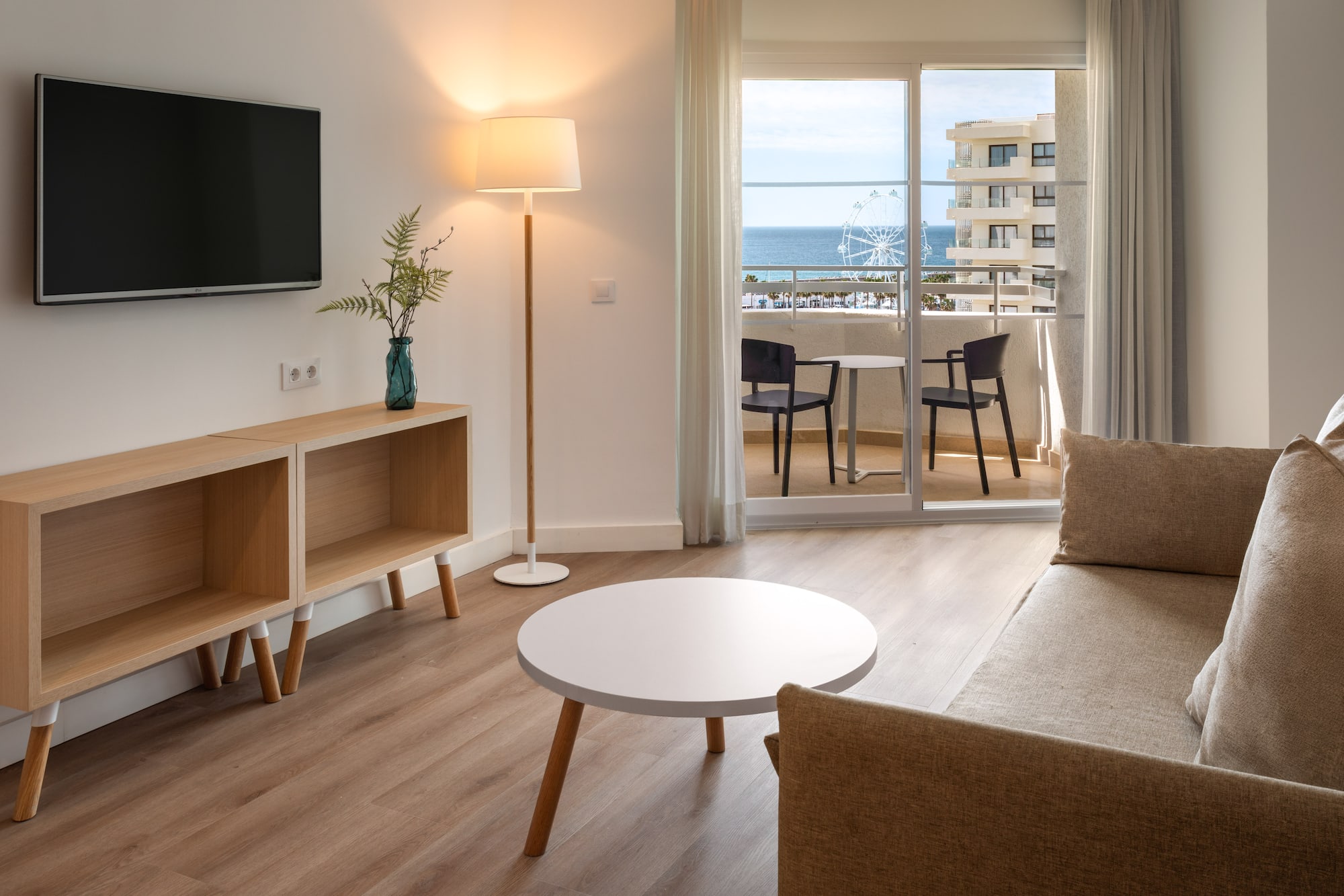 Hotel suite living room with ocean view balcony, TV, sofa, and Ferris wheel.