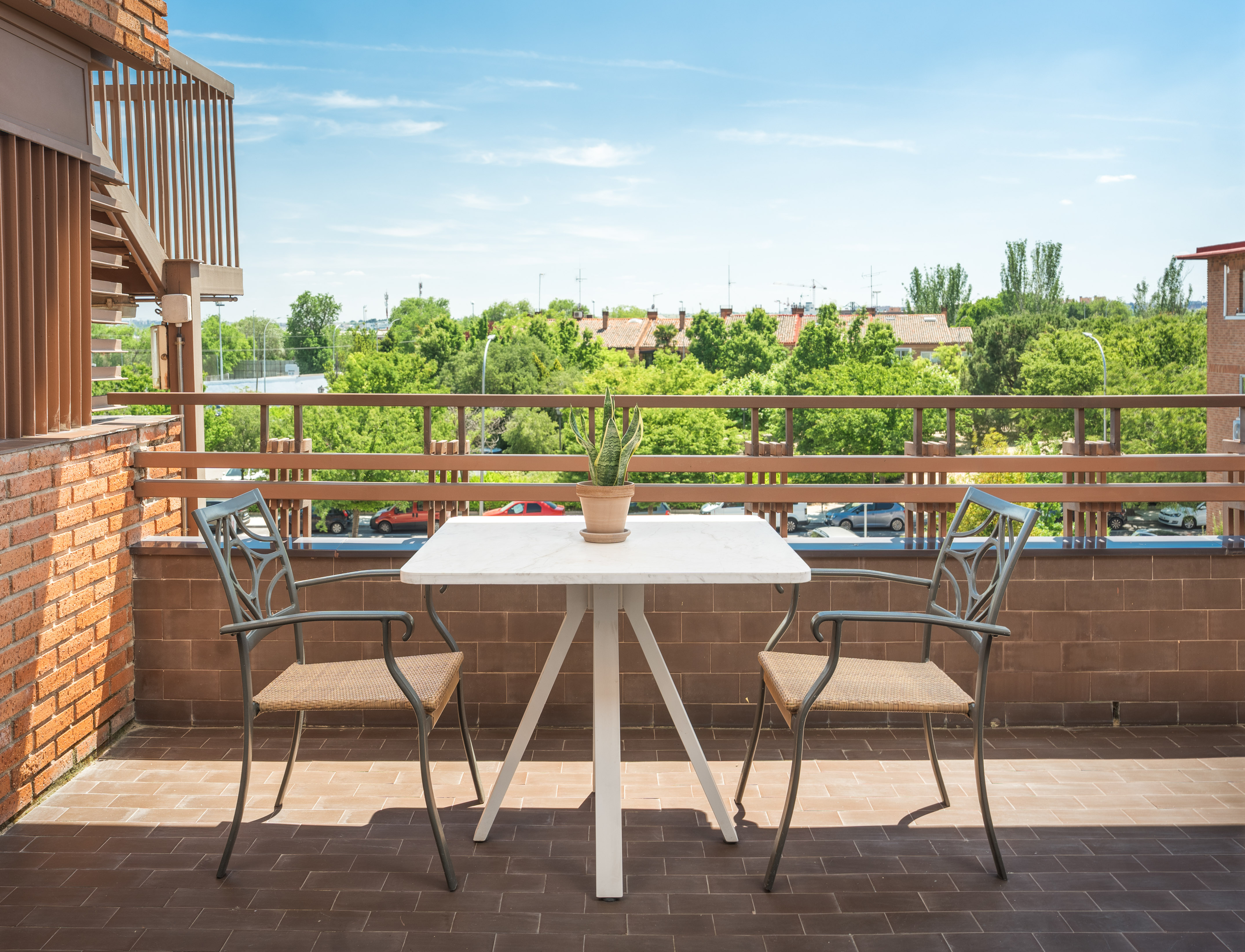 Sunny balcony with table, chairs, potted plant, and green city views.