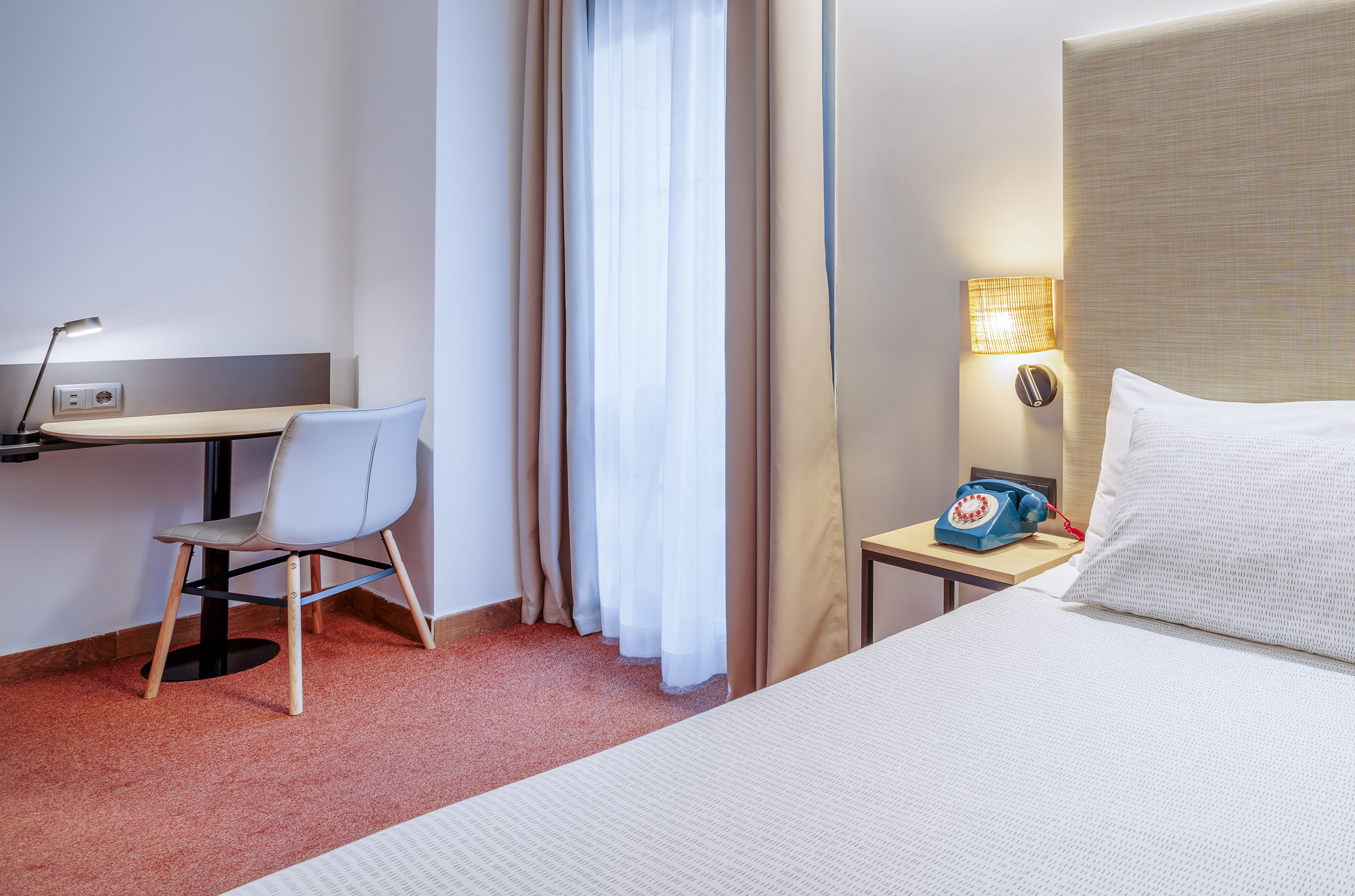 Modern hotel room with work desk, bed, task lamp, and blue vintage phone.
