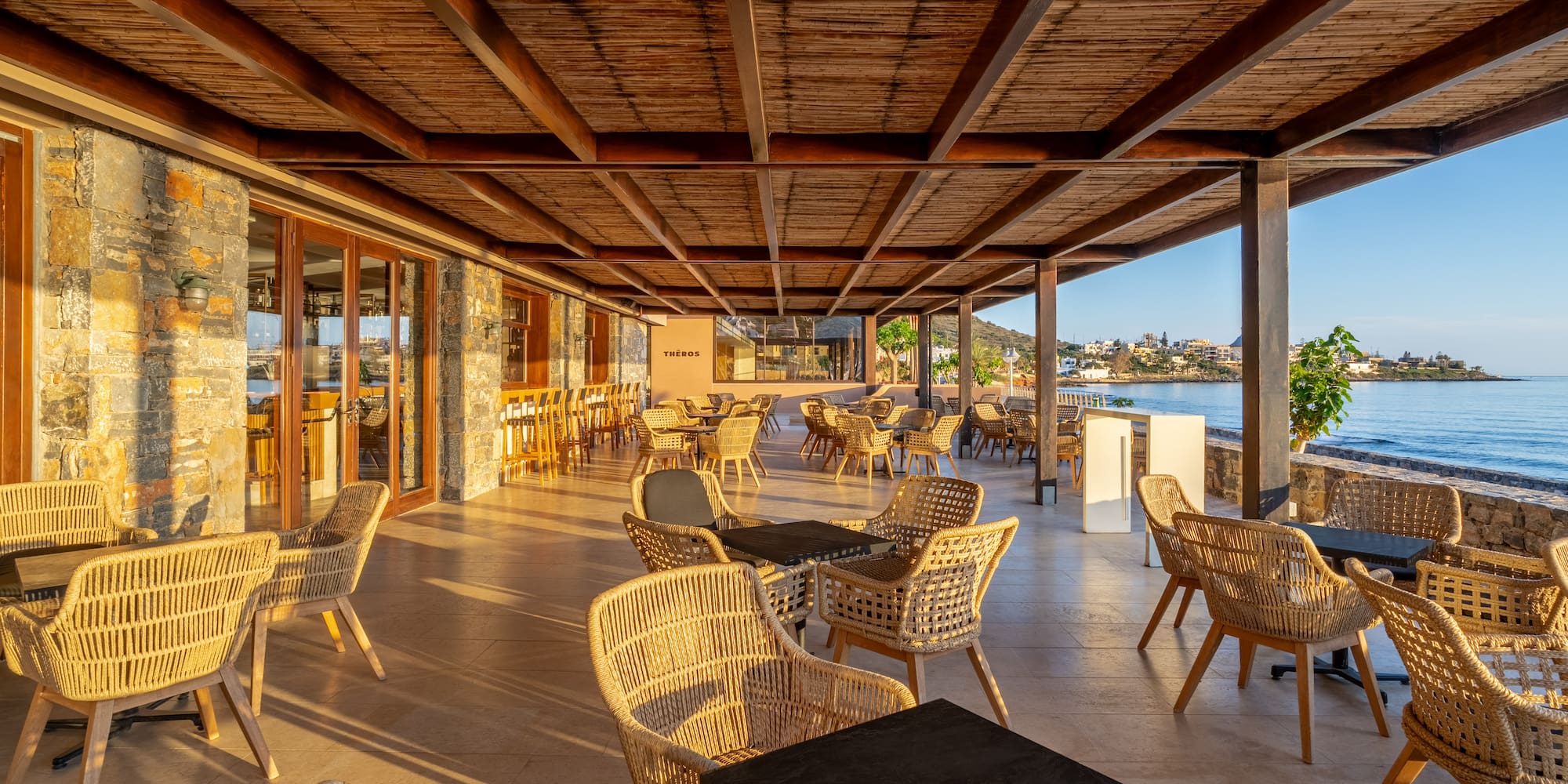 Coastal restaurant patio with woven chairs, wooden ceiling, and sea view.
