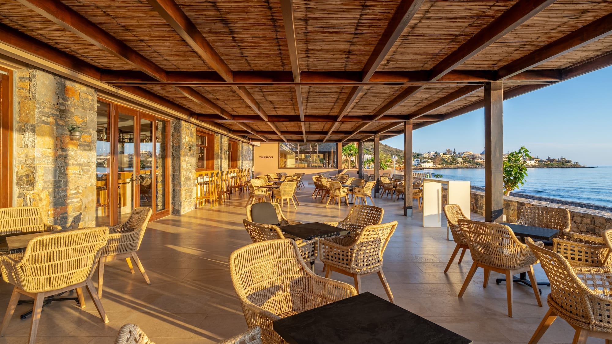 Coastal restaurant patio with woven chairs, wooden ceiling, and sea view.