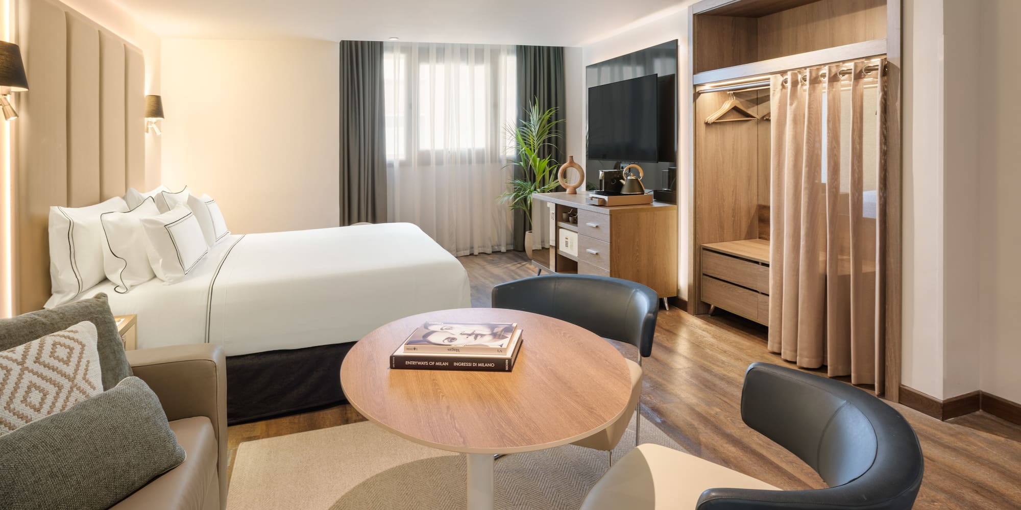 Modern hotel guest room with bed, sofa, TV, and open wardrobe.