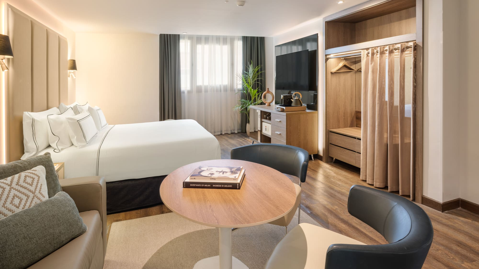 Modern hotel guest room with bed, sofa, TV, and open wardrobe.