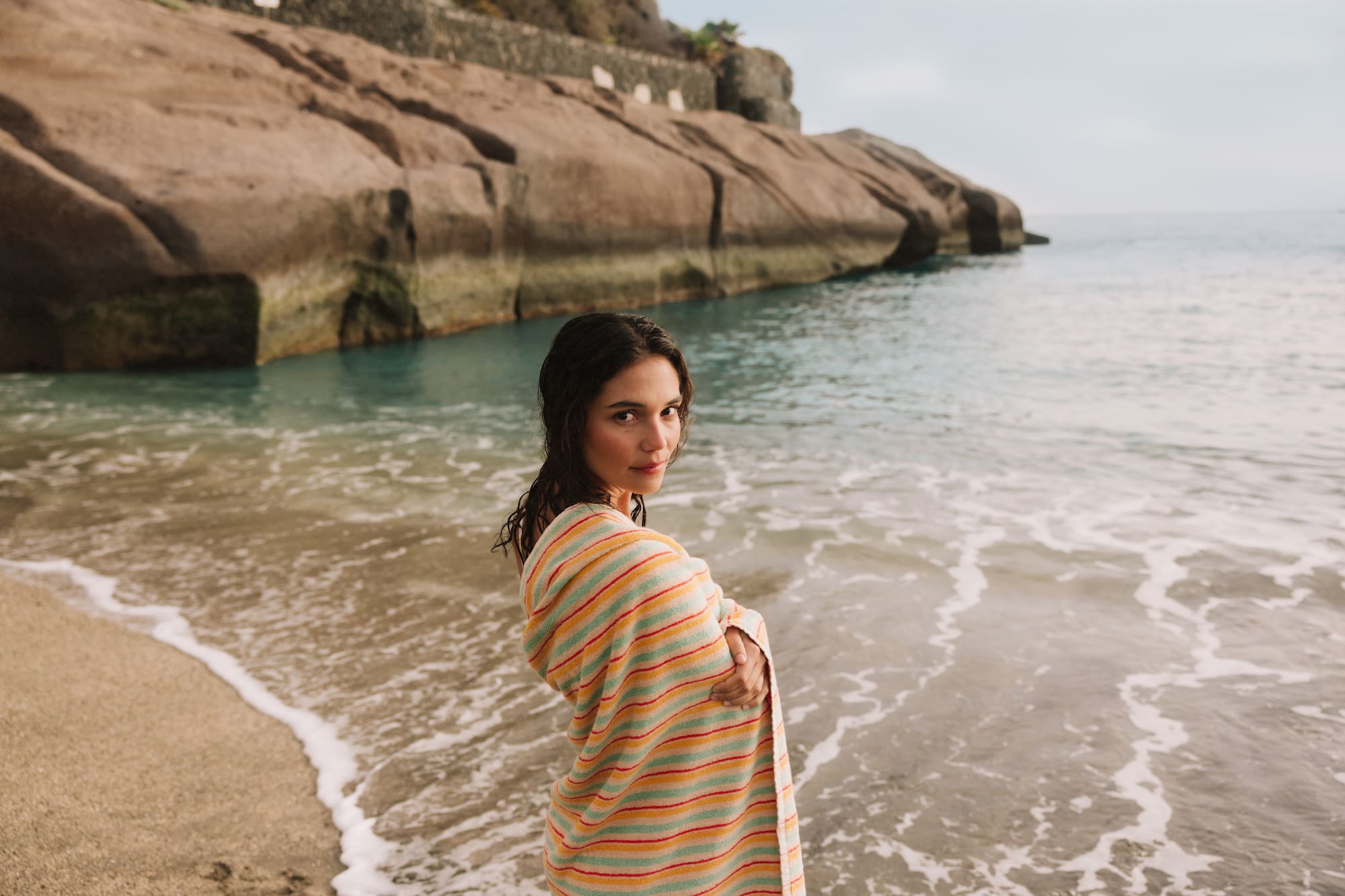 Woman in striped beach towel by ocean, sandy shore, rocky coastline.