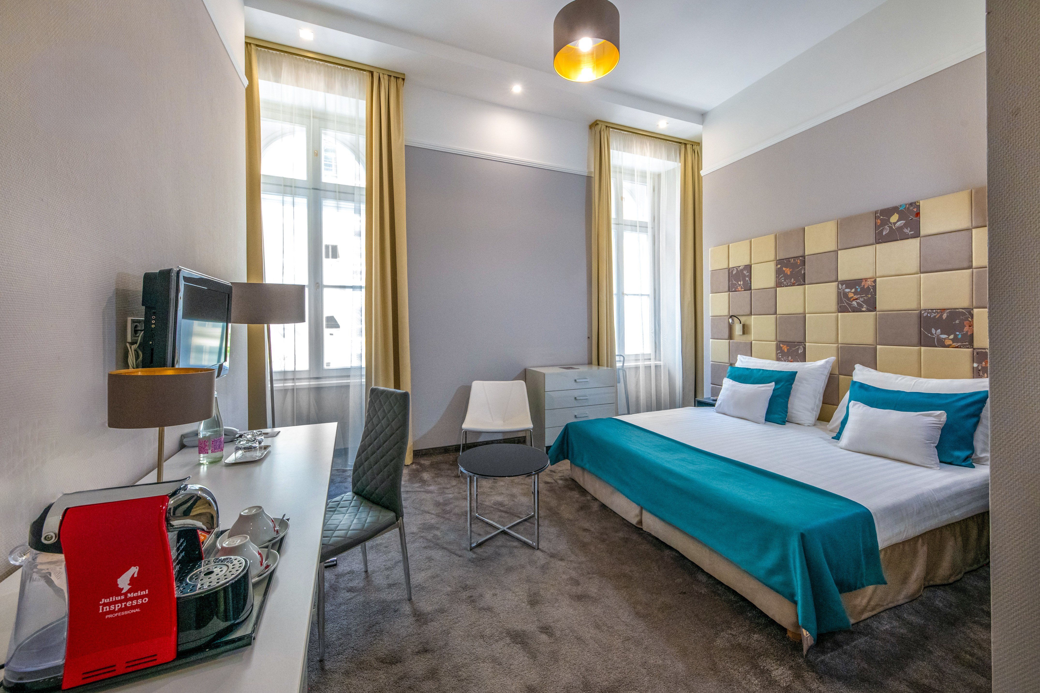 Modern hotel room with teal bedspread, patterned headboard, and coffee maker.