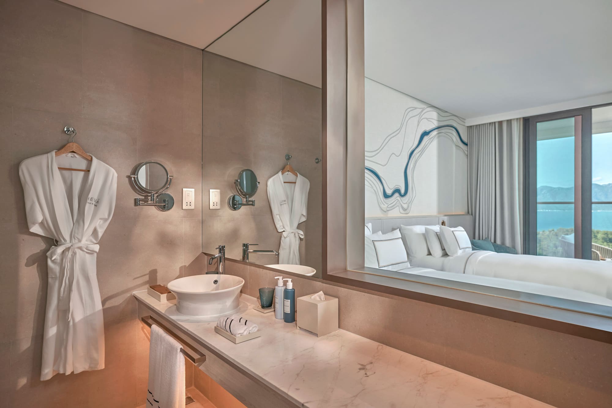 Modern hotel bathroom vanity reflecting bedroom with ocean view.