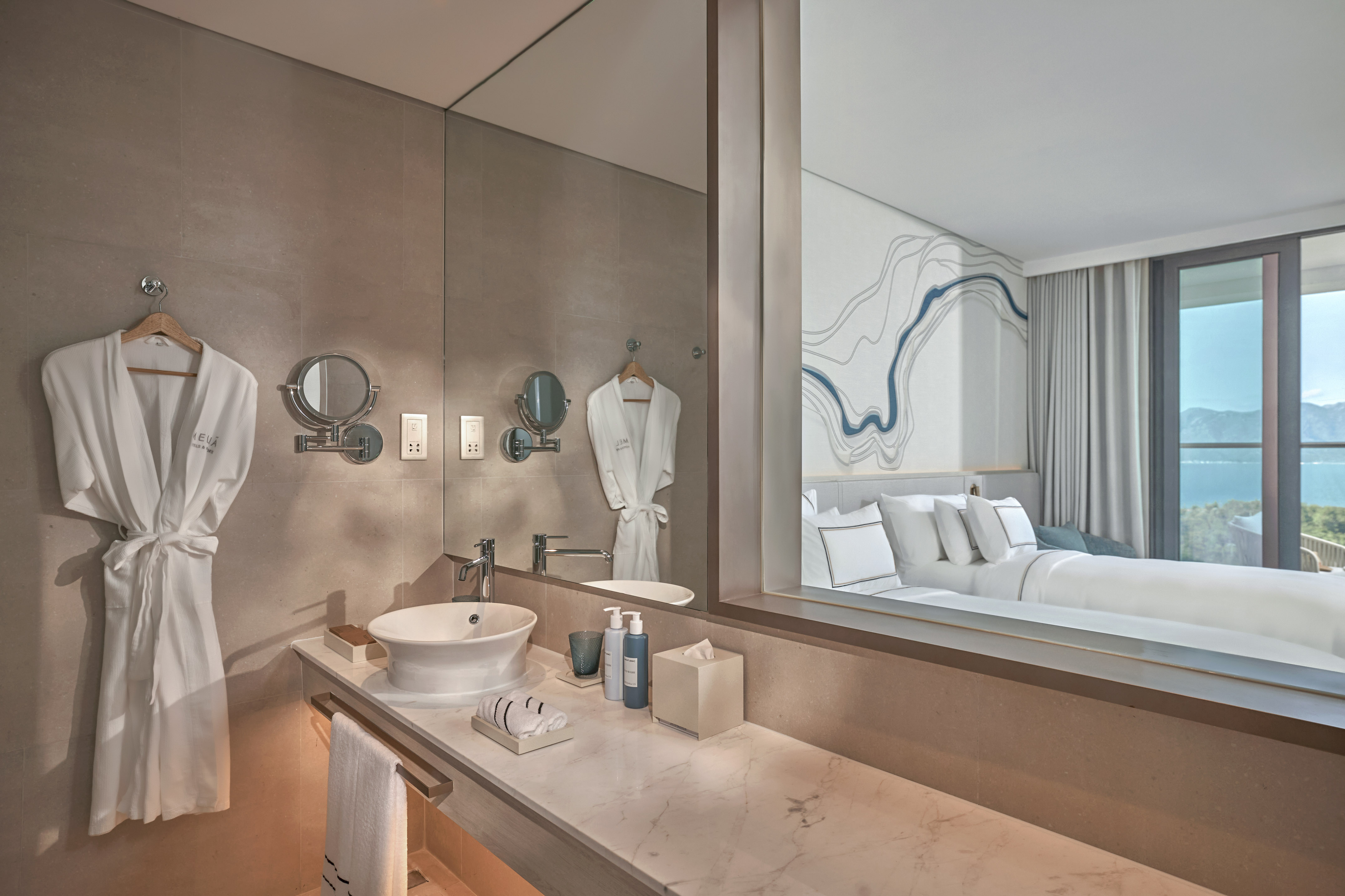 Modern hotel bathroom vanity reflecting bedroom with ocean view.