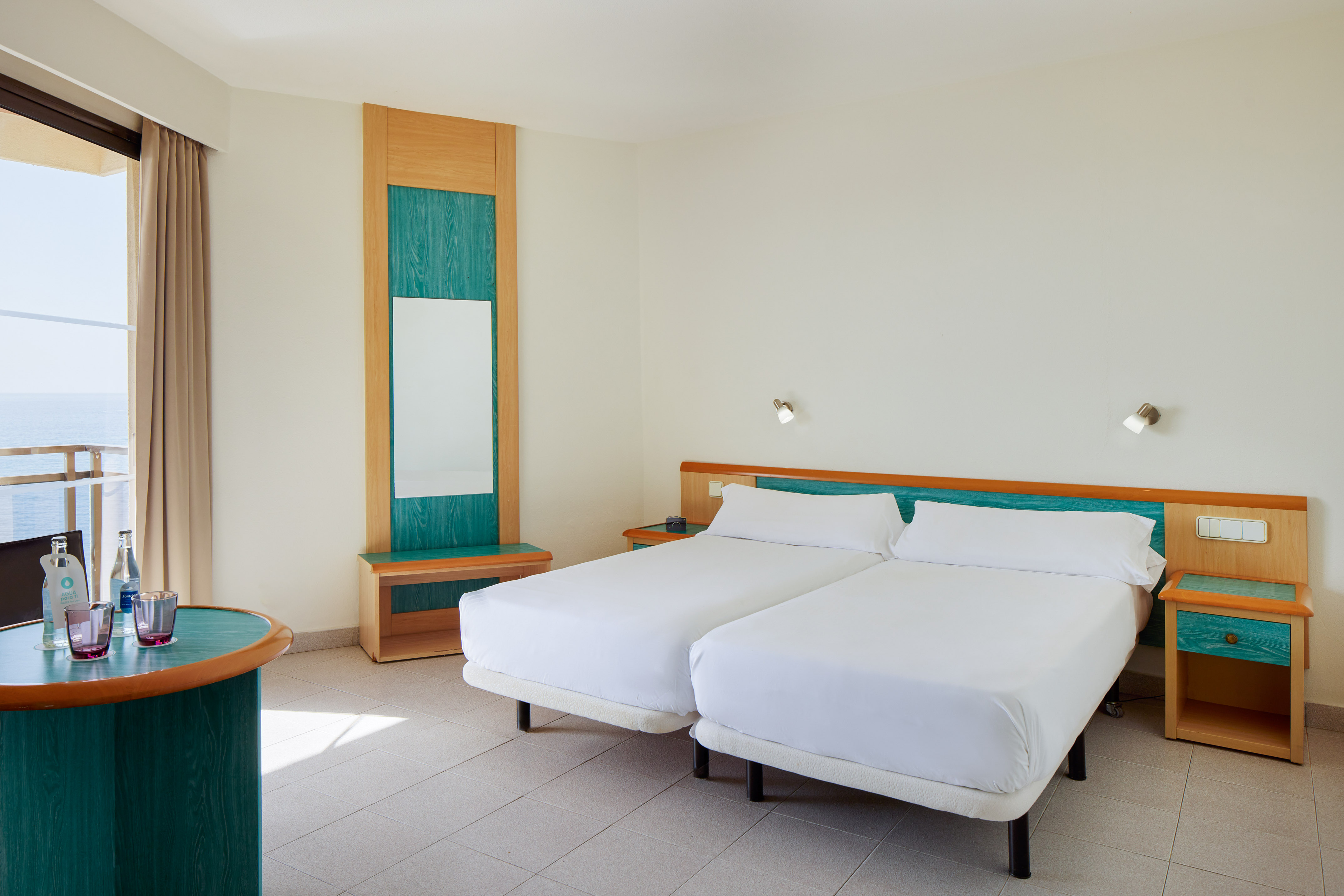 Bright resort room with twin beds, teal accents, and ocean view balcony.