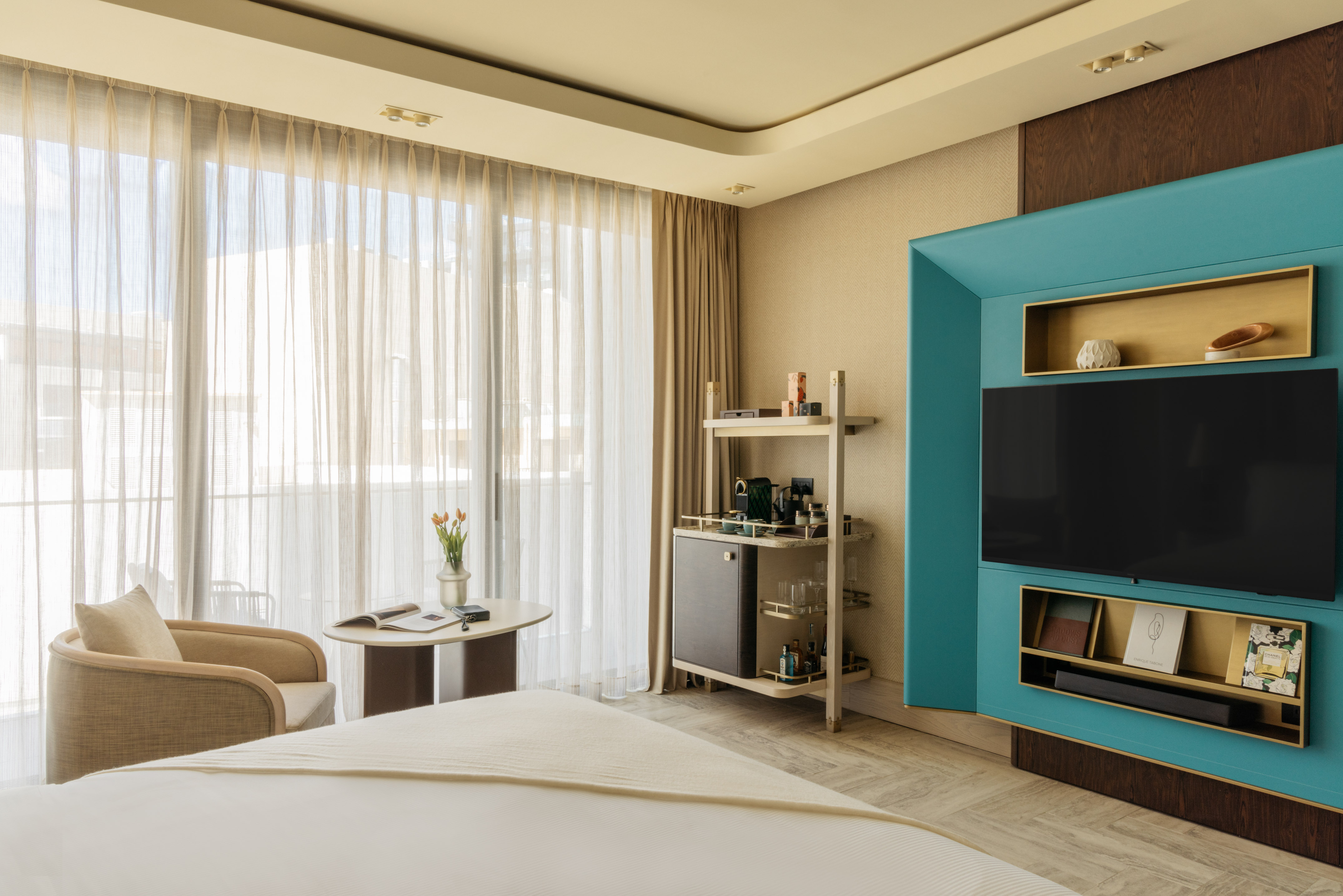 Modern hotel bedroom with large window, teal TV unit, and minibar.