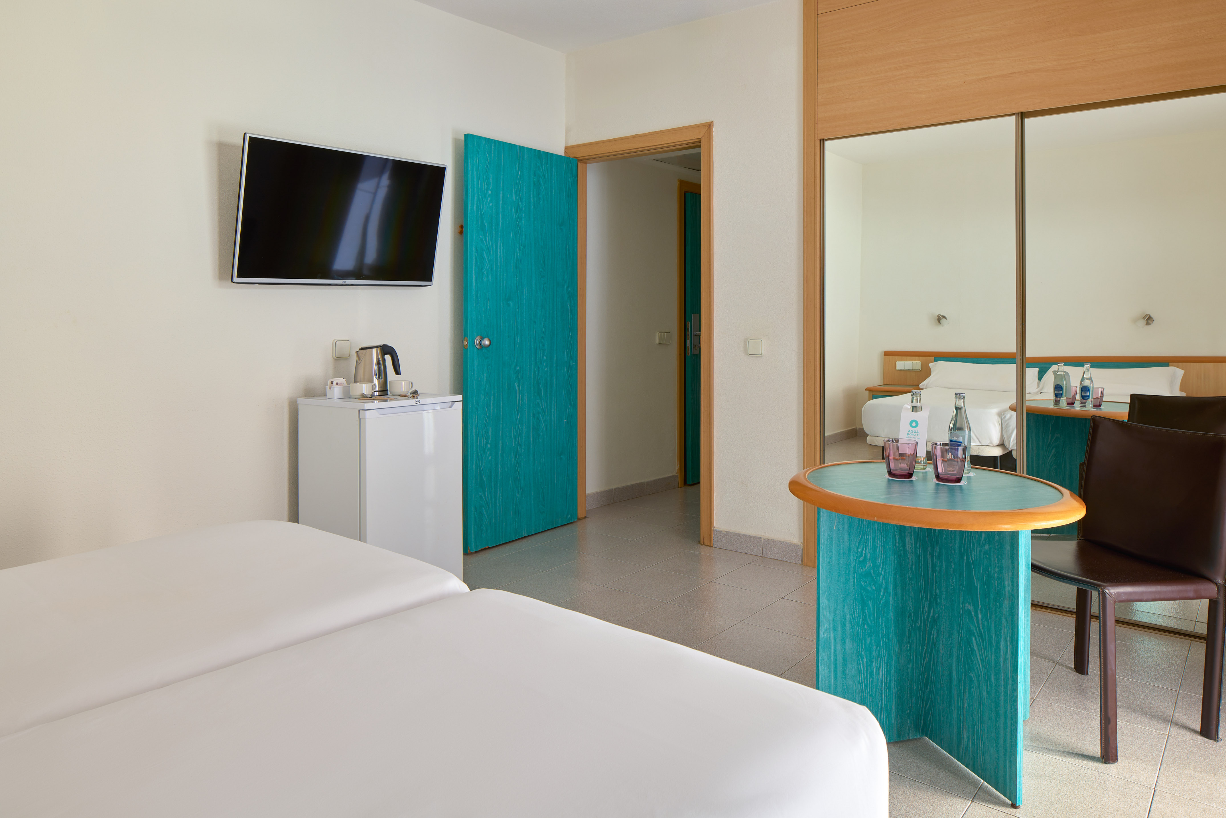 Modern hotel room with twin beds, TV, mini-fridge, and teal accents.