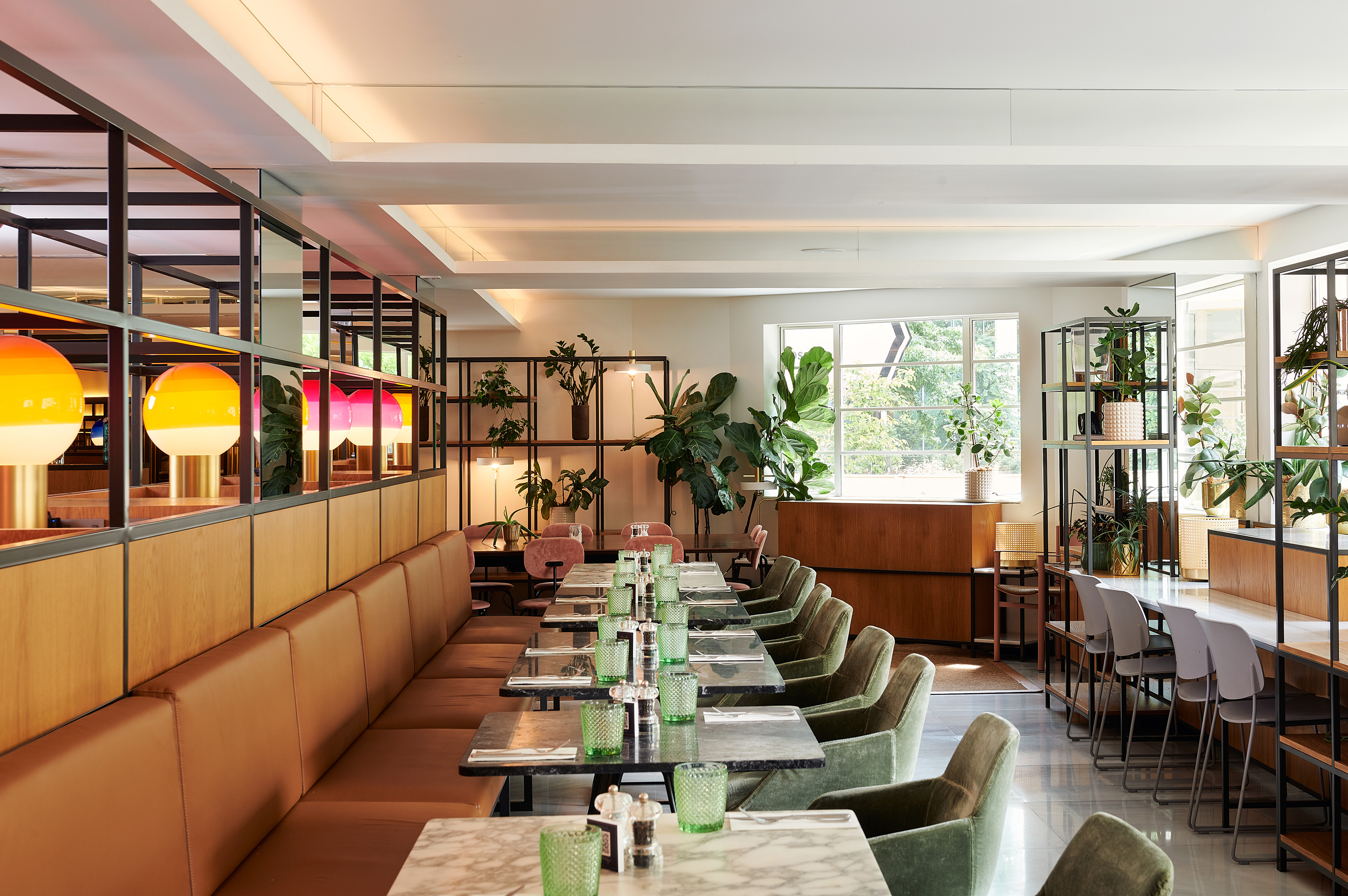 Modern restaurant with brown booths, green chairs, marble tables, and lush plants.