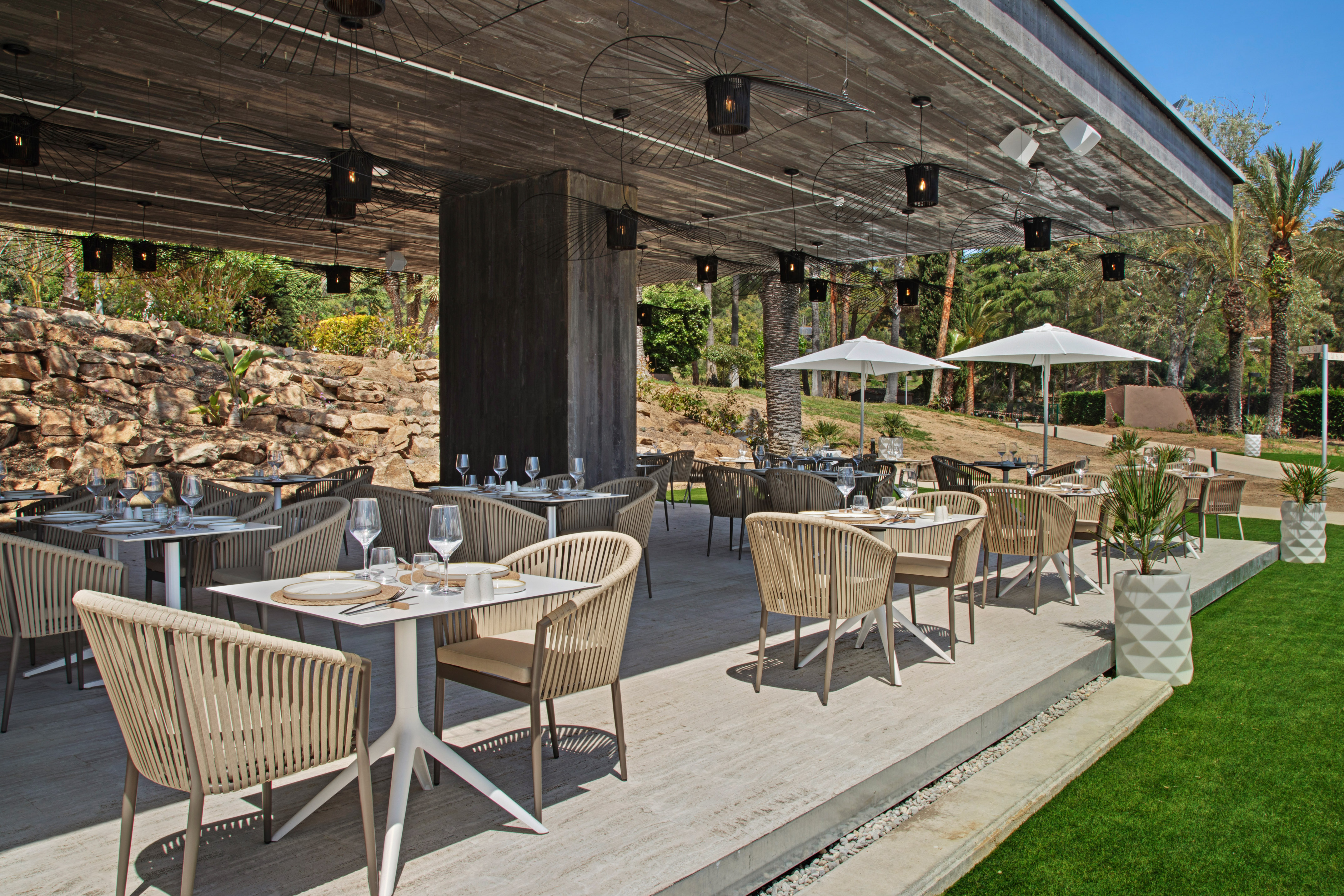 Modern outdoor dining patio with wicker chairs, set tables, and lush garden.