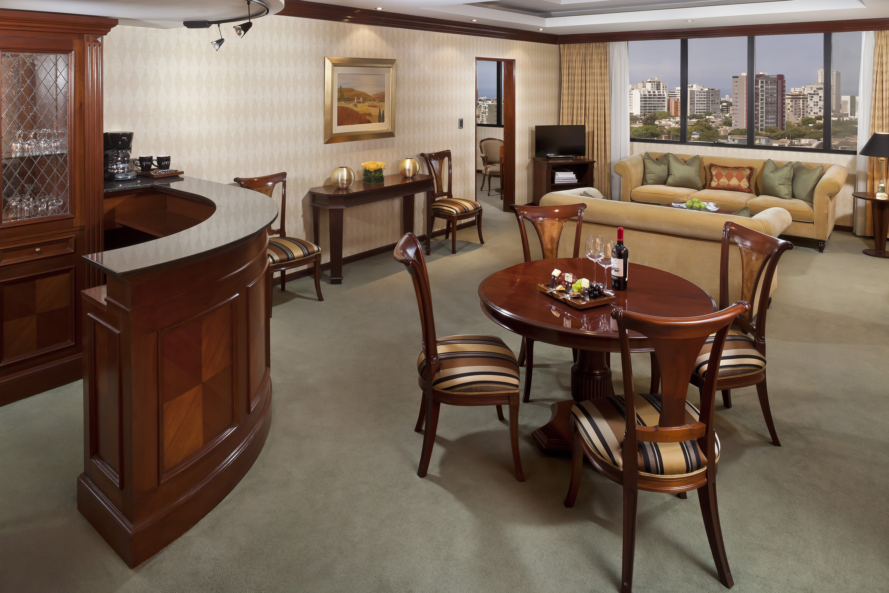 Elegant hotel suite with bar, dining table, sofas, and city skyline view.