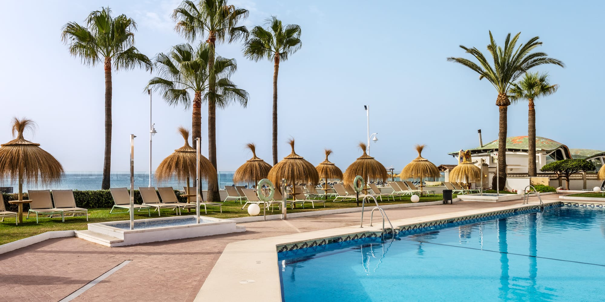 Tropical resort pool with palm trees, thatched umbrellas, sunbeds, and ocean view.