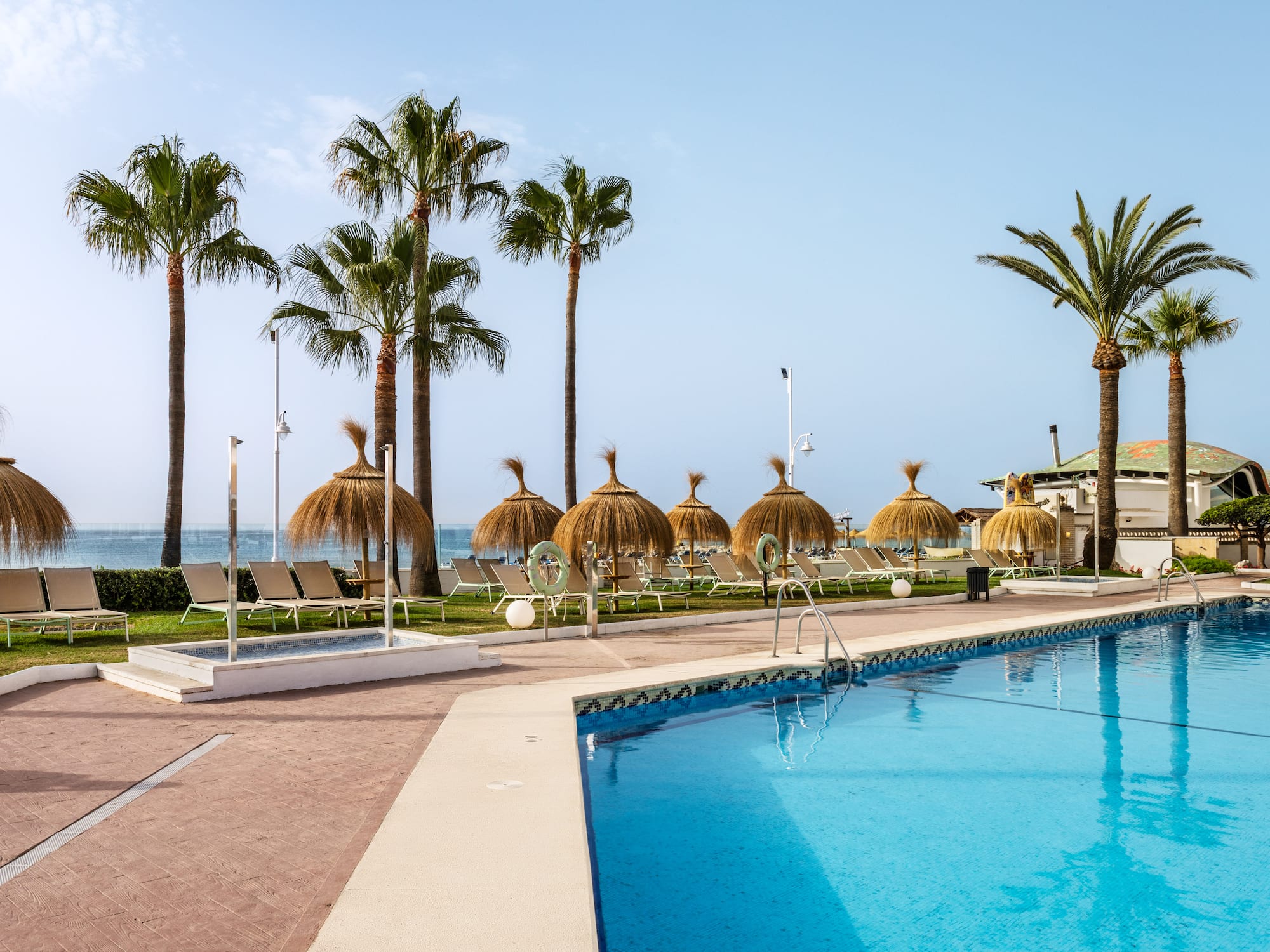 Resort swimming pool, palm trees, thatched parasols, lounge chairs, ocean view.