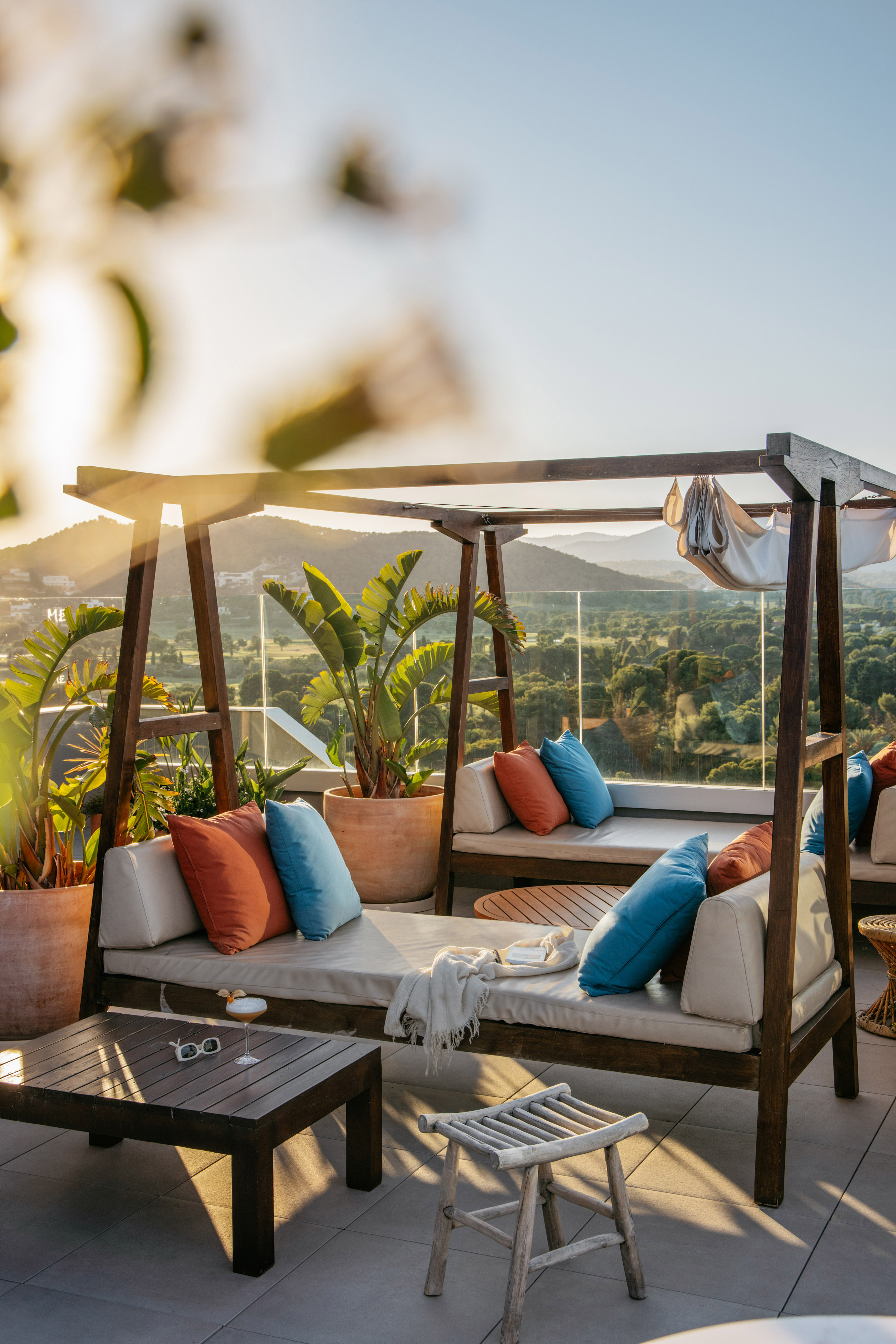 Rooftop lounge with daybeds, colorful cushions, sunset over scenic mountains.