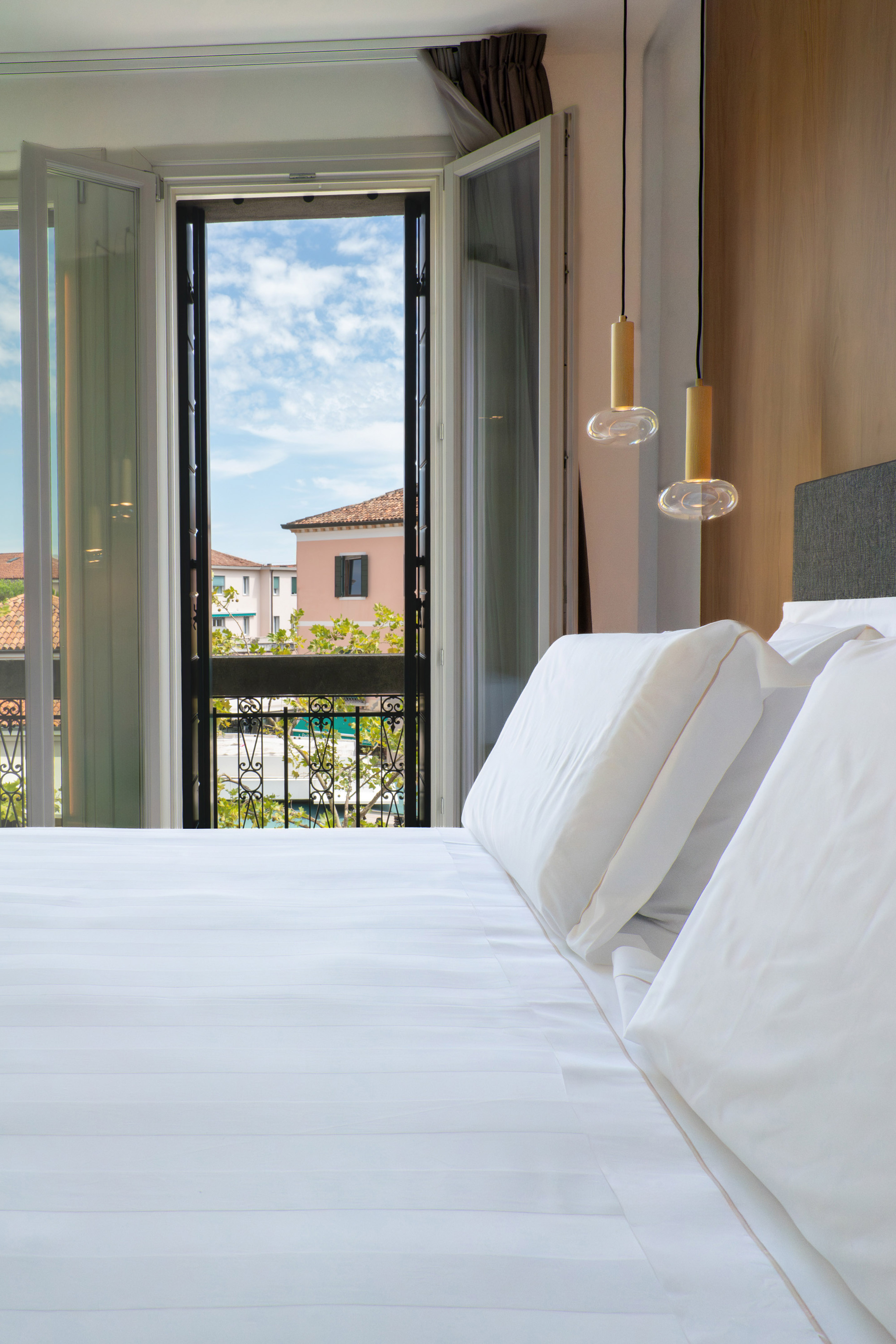 Modern bedroom with white bed, pendant lights, and open balcony overlooking buildings.