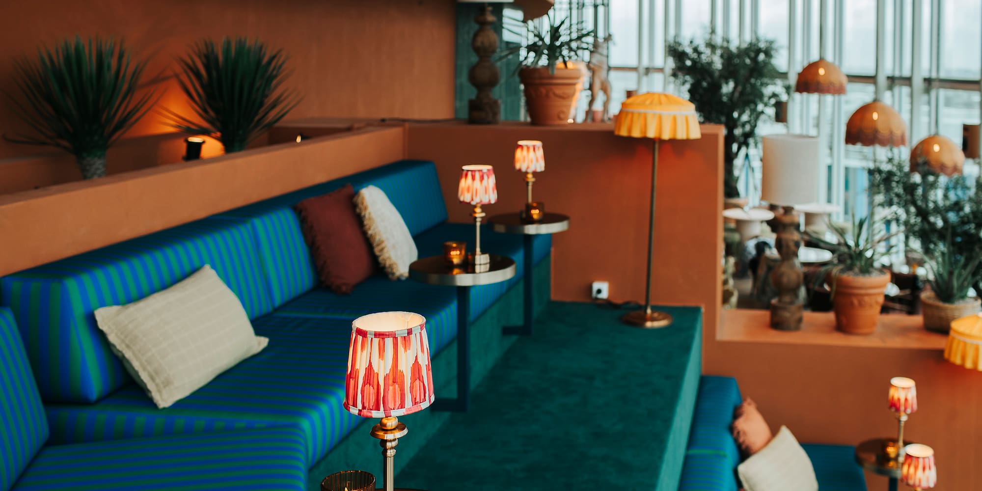 Modern lounge with blue-green striped seating, terracotta walls, decorative lamps, and plants.