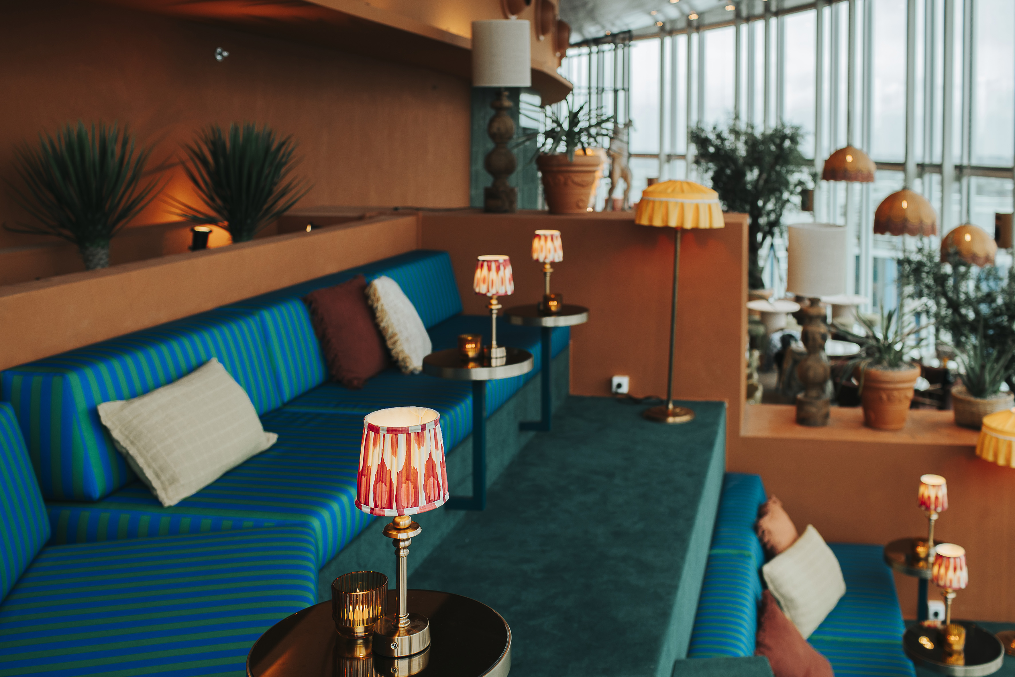 Modern lounge with blue-green striped seating, terracotta walls, decorative lamps, and plants.
