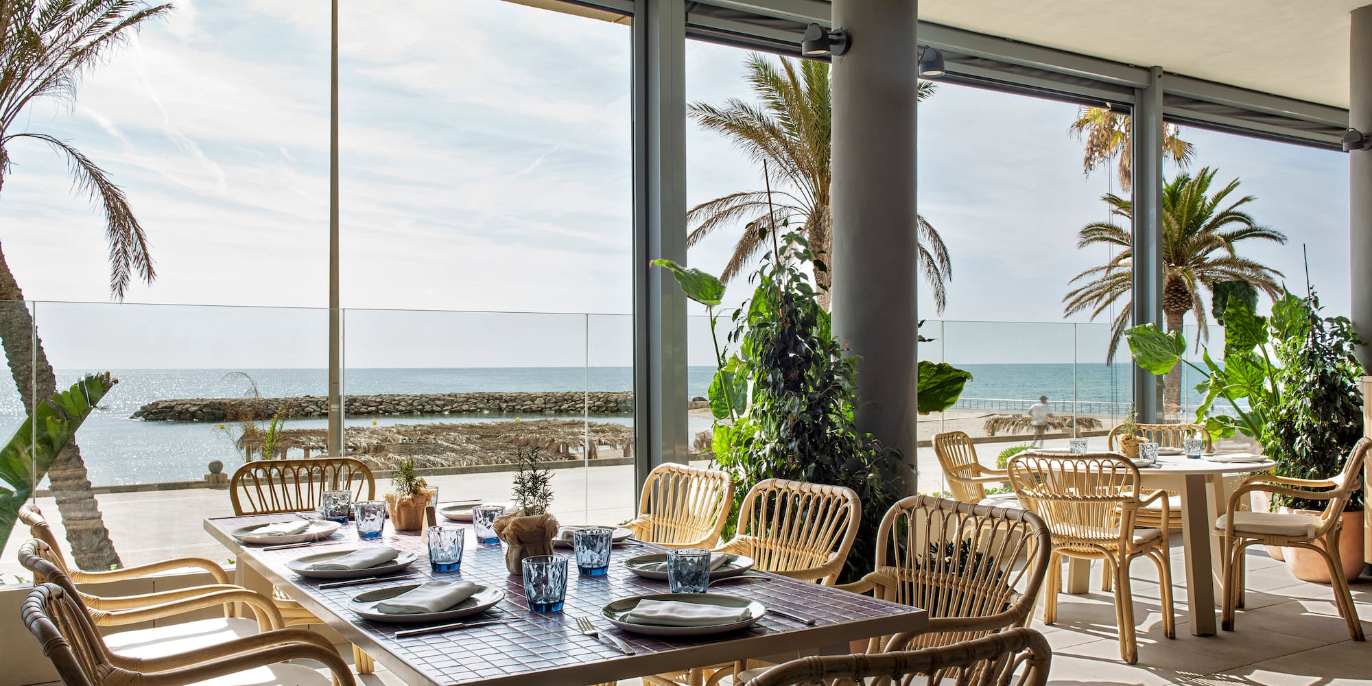 Beachfront restaurant dining area with ocean view, palm trees, and rattan chairs.