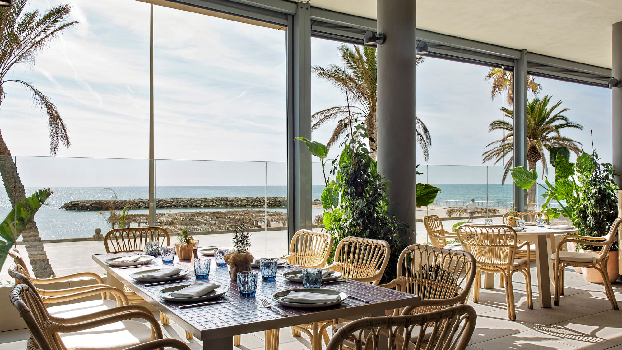 Beachfront restaurant dining area with ocean view, palm trees, and rattan chairs.