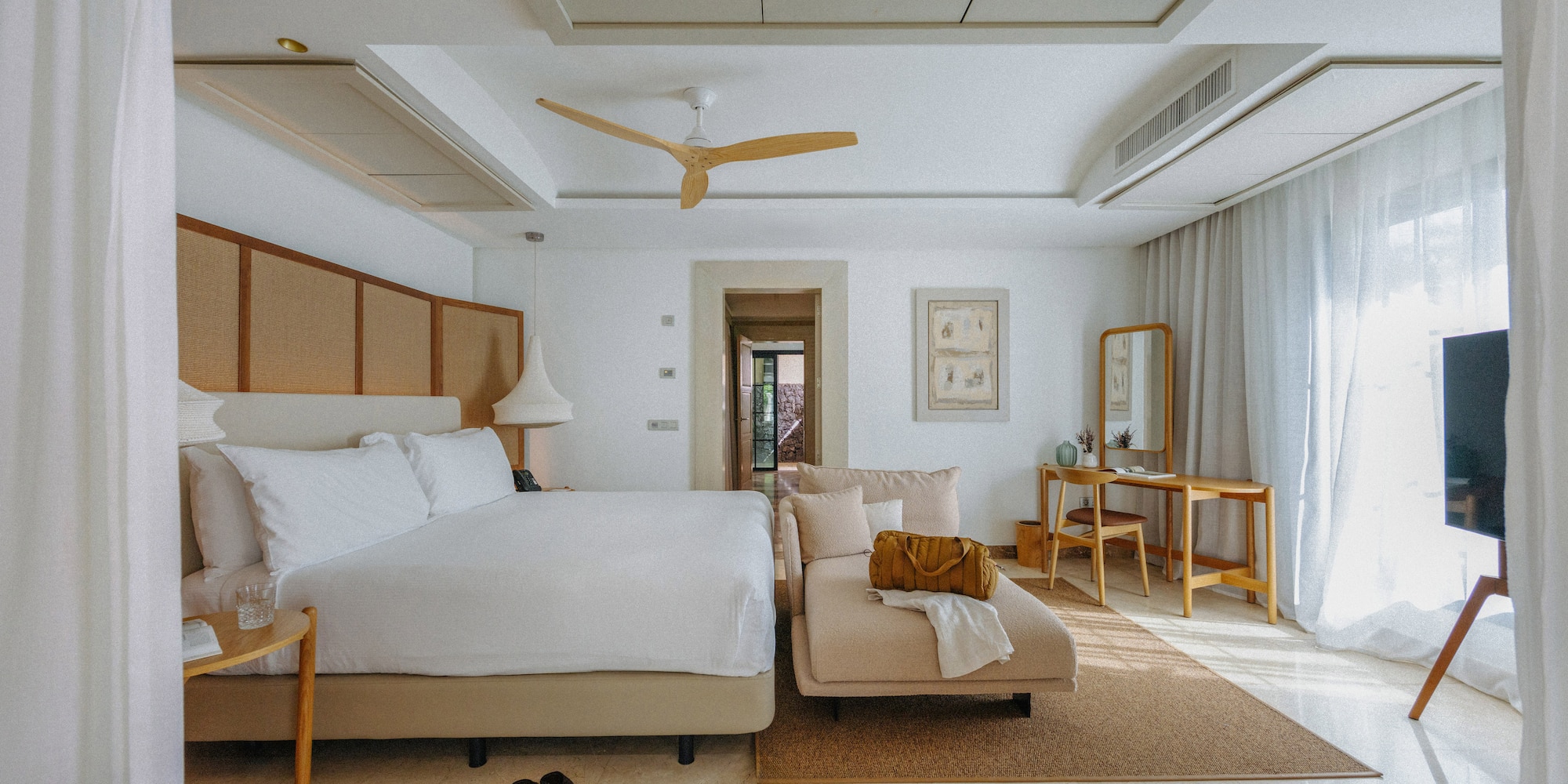 Spacious modern hotel bedroom with white bed, chaise lounge, and wood fan.
