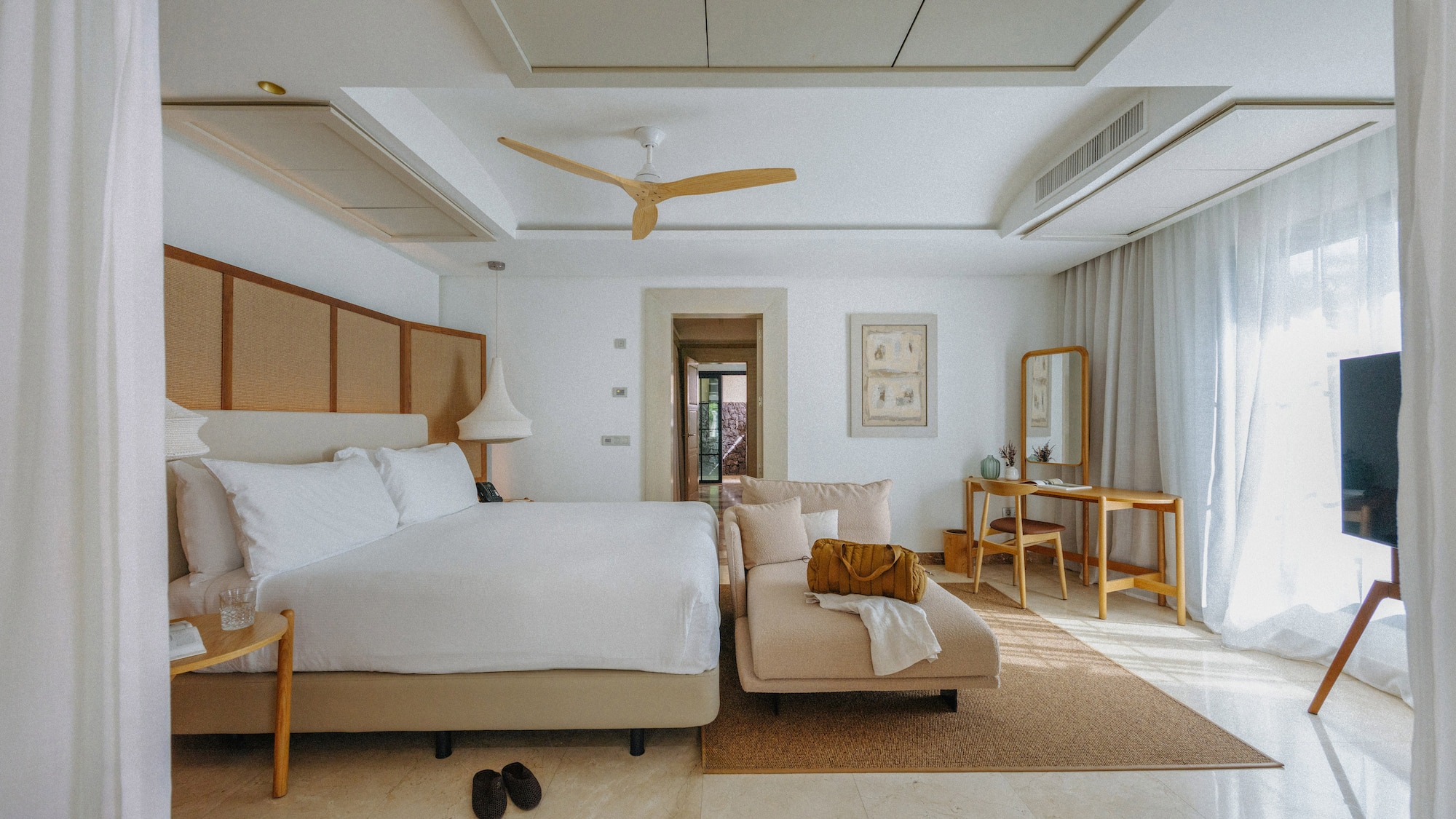 Spacious modern hotel bedroom with white bed, chaise lounge, and wood fan.