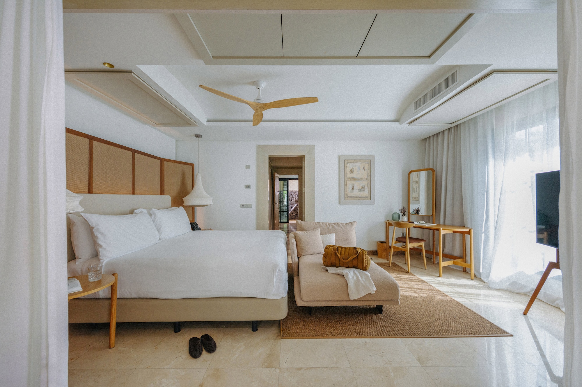 Spacious modern hotel bedroom with white bed, chaise lounge, and wood fan.