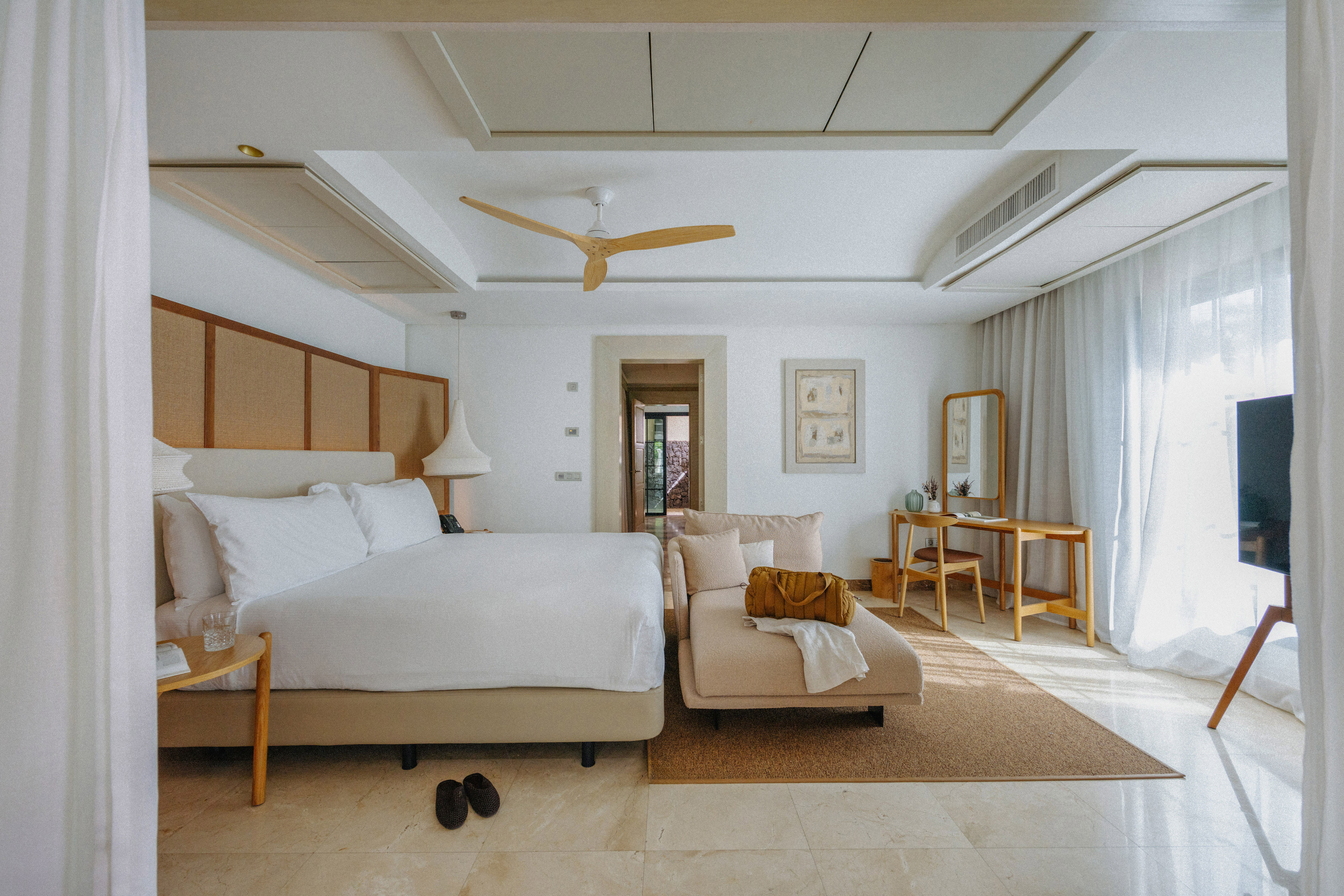Spacious modern hotel bedroom with white bed, chaise lounge, and wood fan.