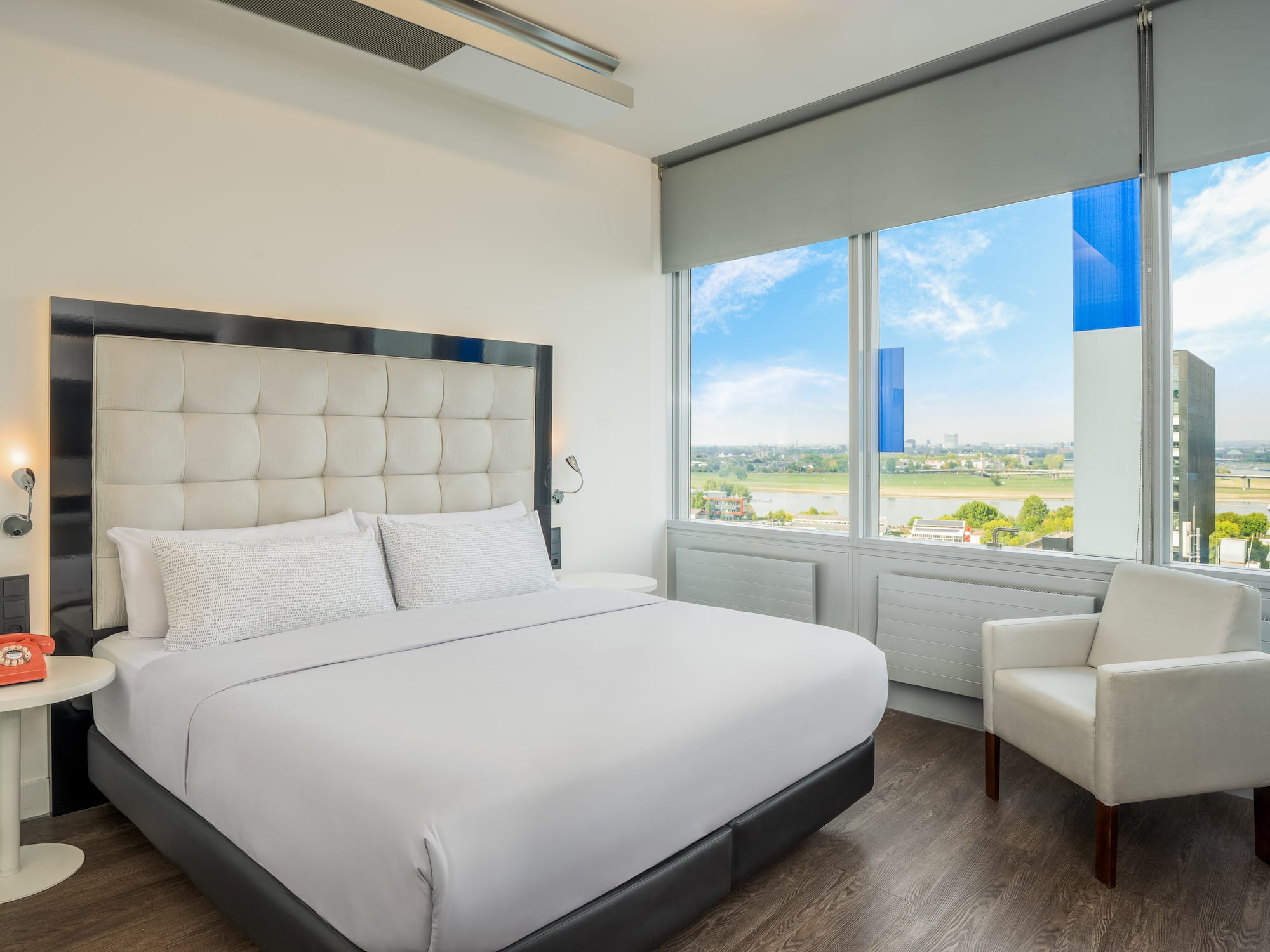 Bright hotel room with white bed, city view, and red vintage telephone.