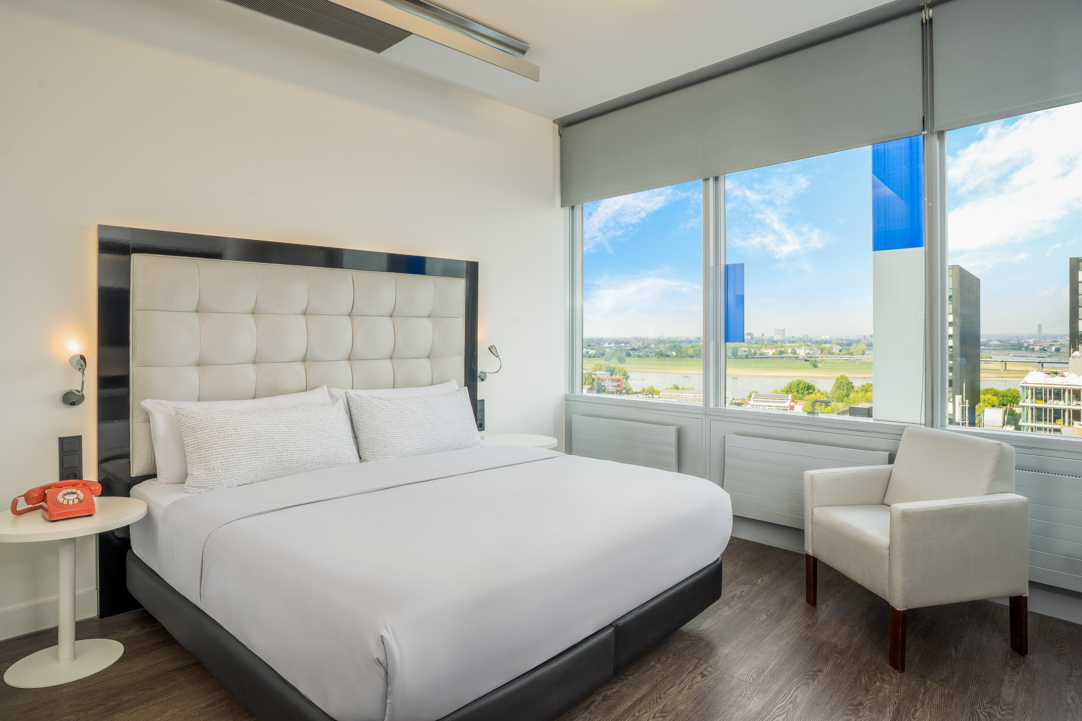Bright hotel room with white bed, city view, and red vintage telephone.