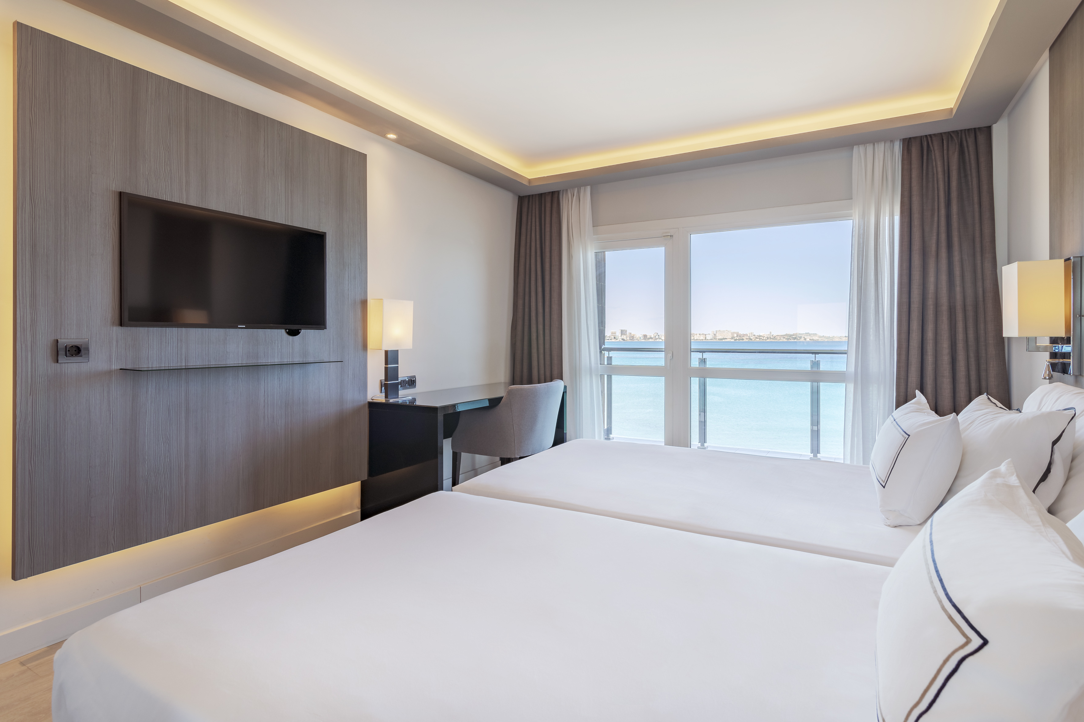 Modern hotel room with twin beds, sea view, TV, and desk by large window.