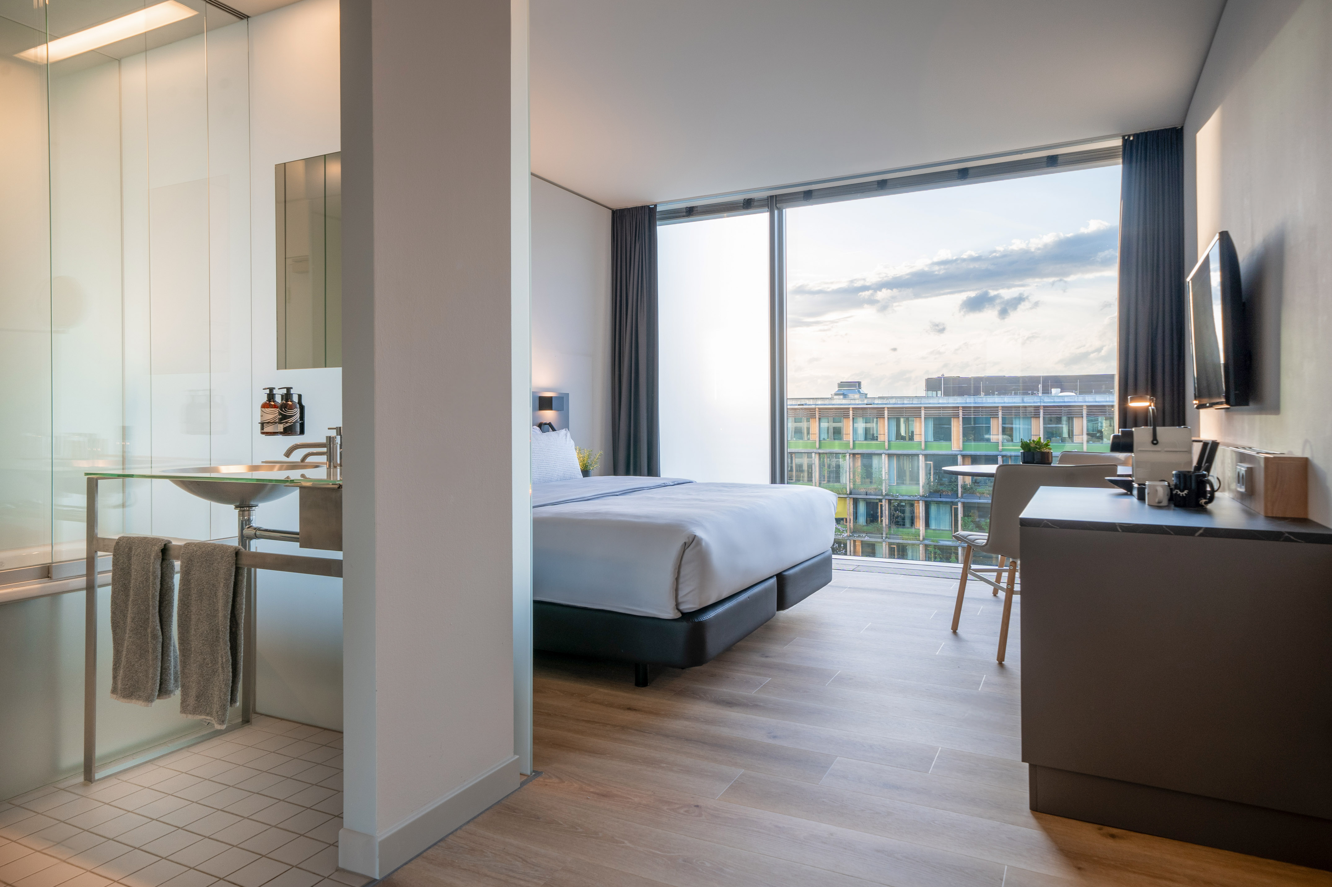 Contemporary hotel suite with king bed, open bathroom, desk, and city view.