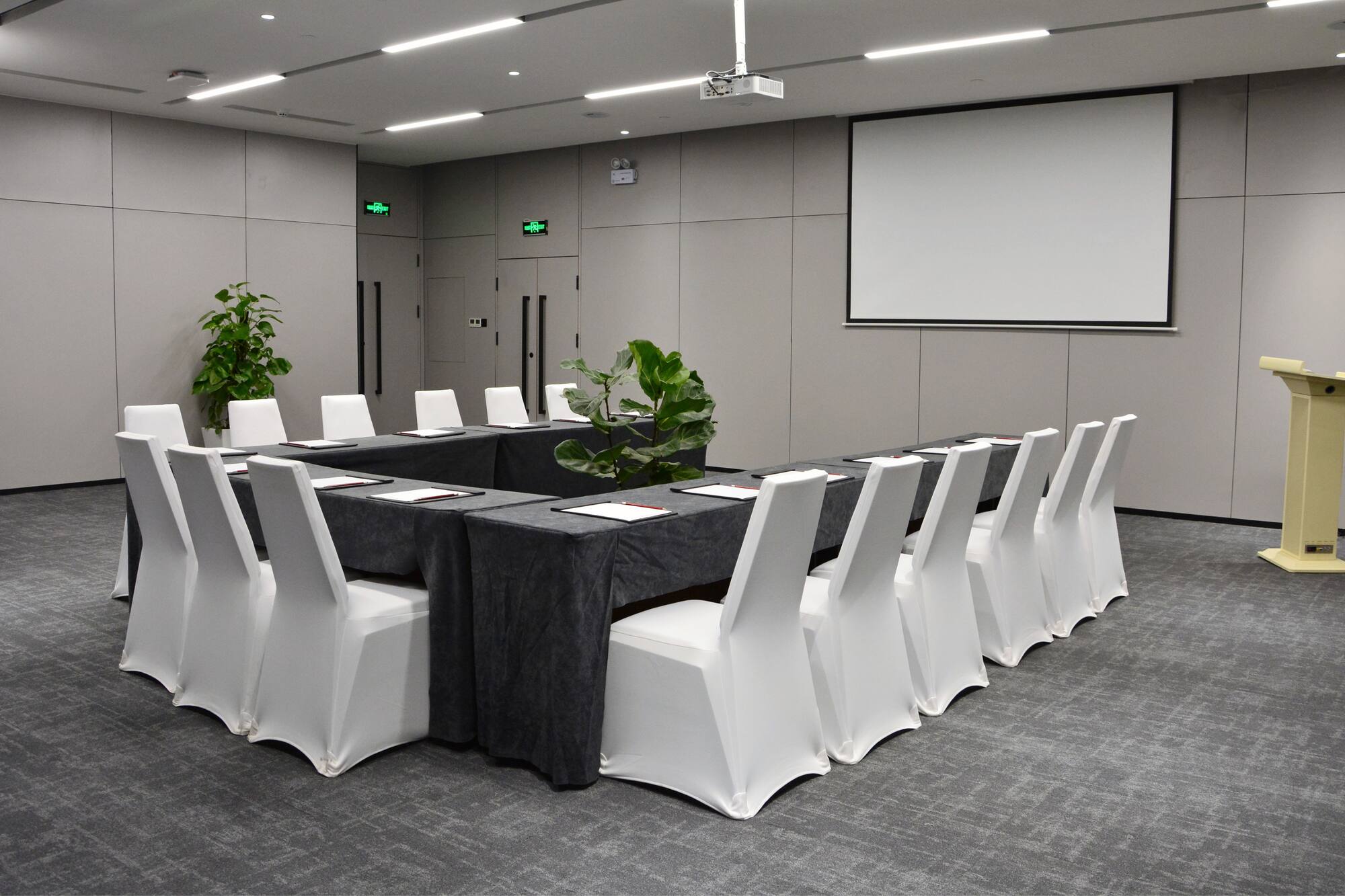 Conference room with U-shaped table, white chairs, projector screen, lectern.