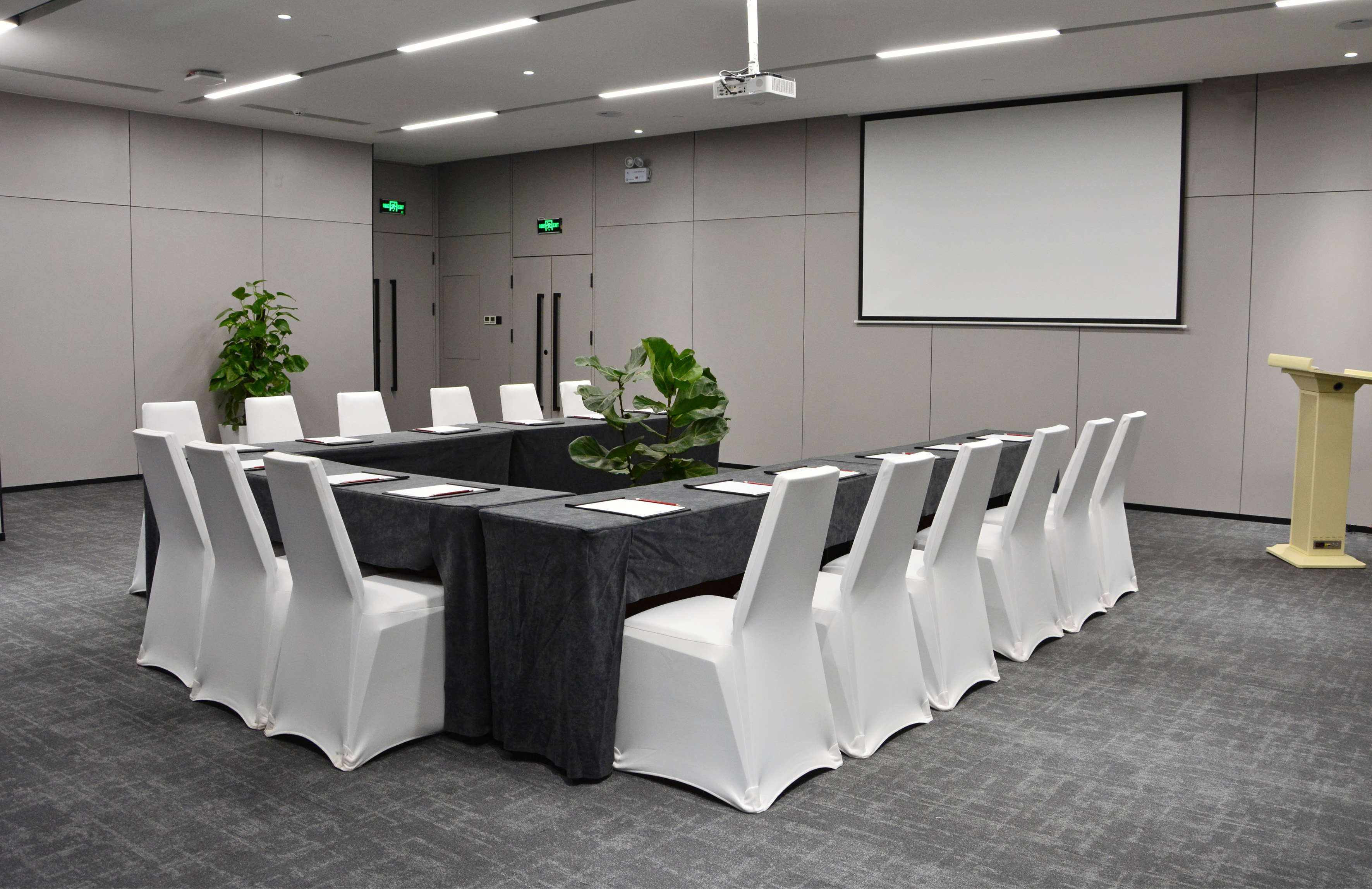 Conference room with U-shaped table, white chairs, projector screen, lectern.