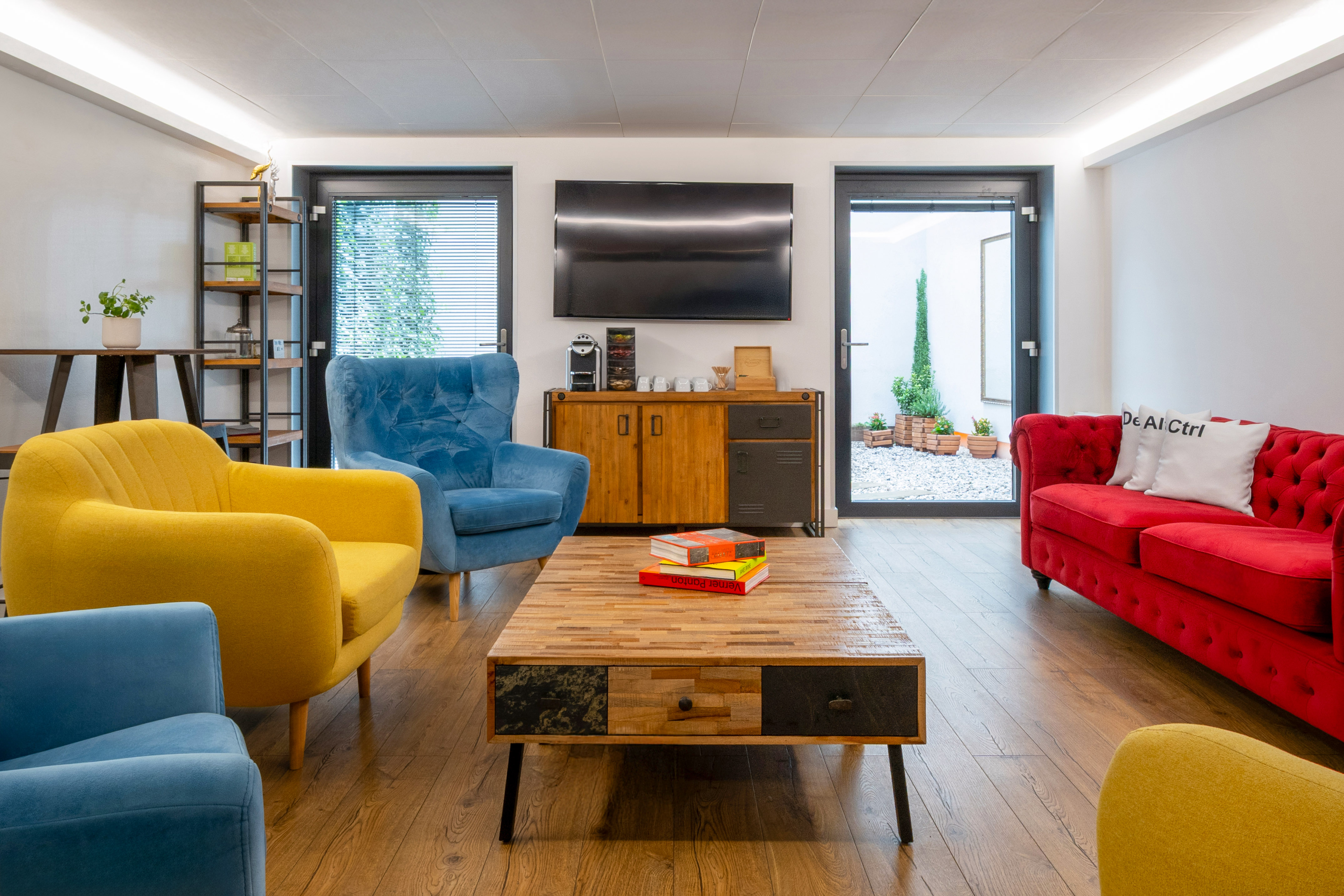 Modern colorful lounge area with red sofa, blue and yellow armchairs, wood table, and TV.