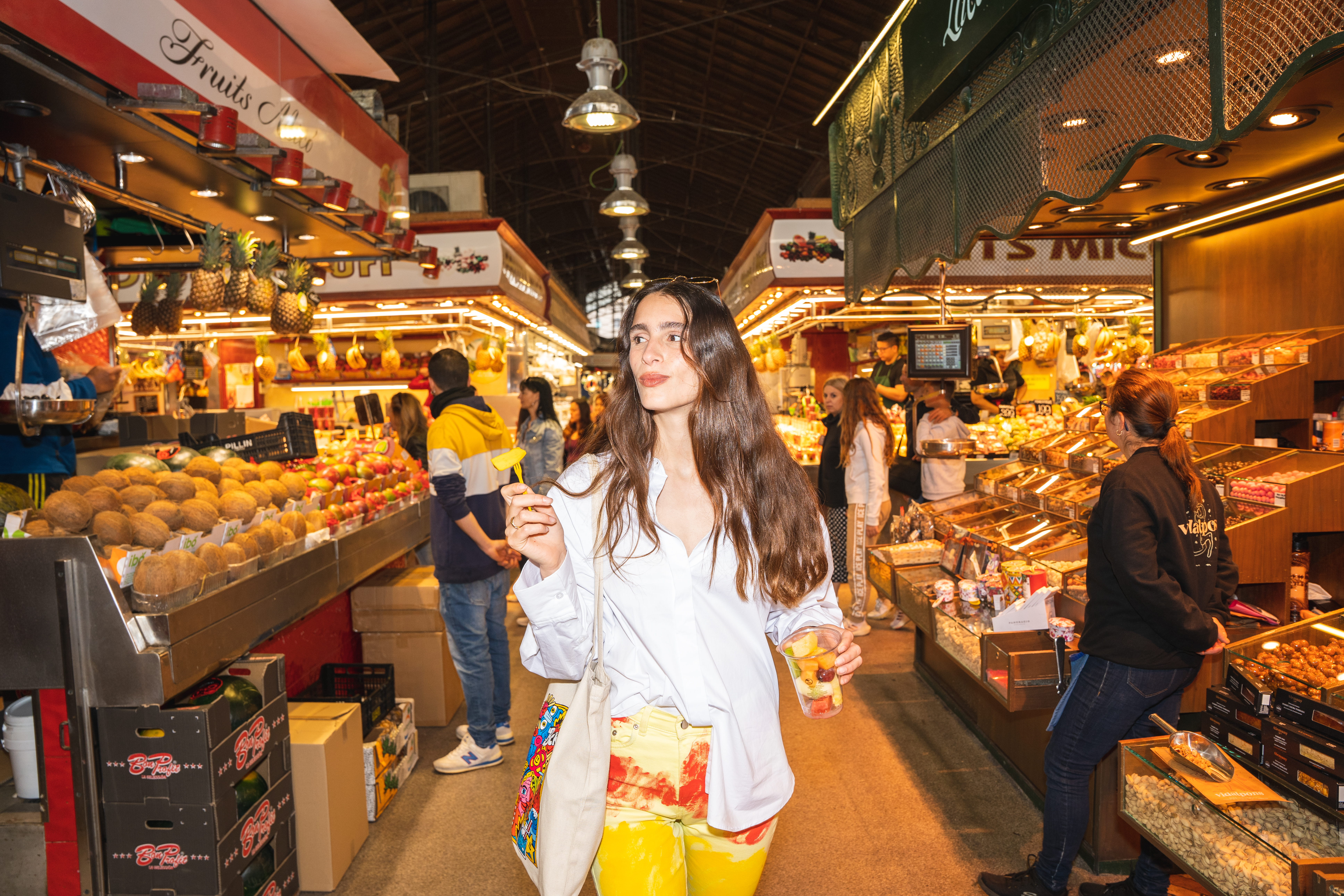Mujer con copa de fruta paseando por un vibrante mercado de abastos.