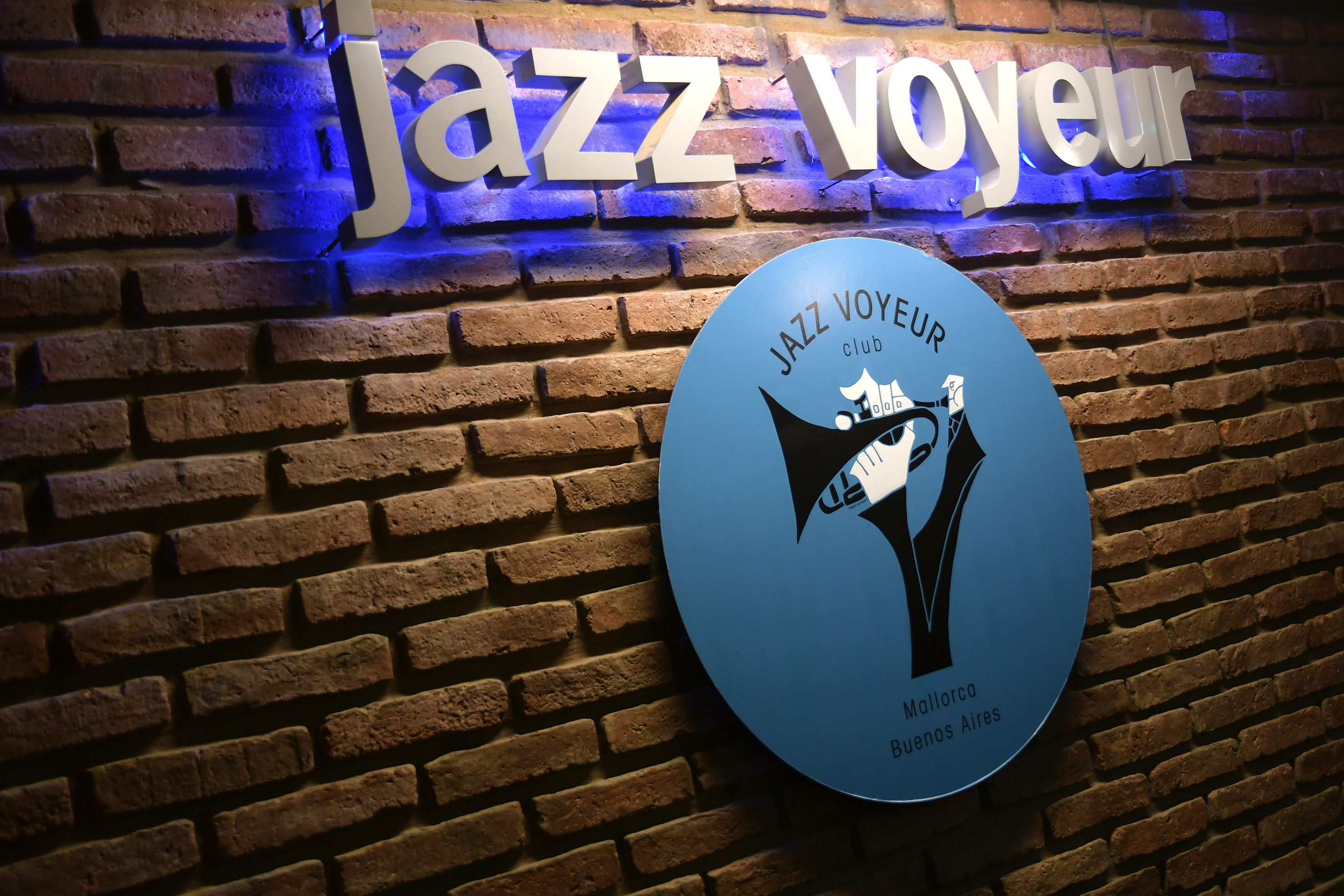 Backlit 'Jazz Voyeur' sign and blue club logo with trumpet on a brick wall.