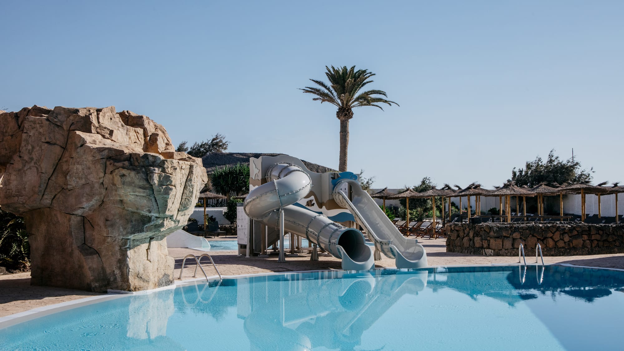 Resort swimming pool with water slides, palm tree, and thatched parasols.